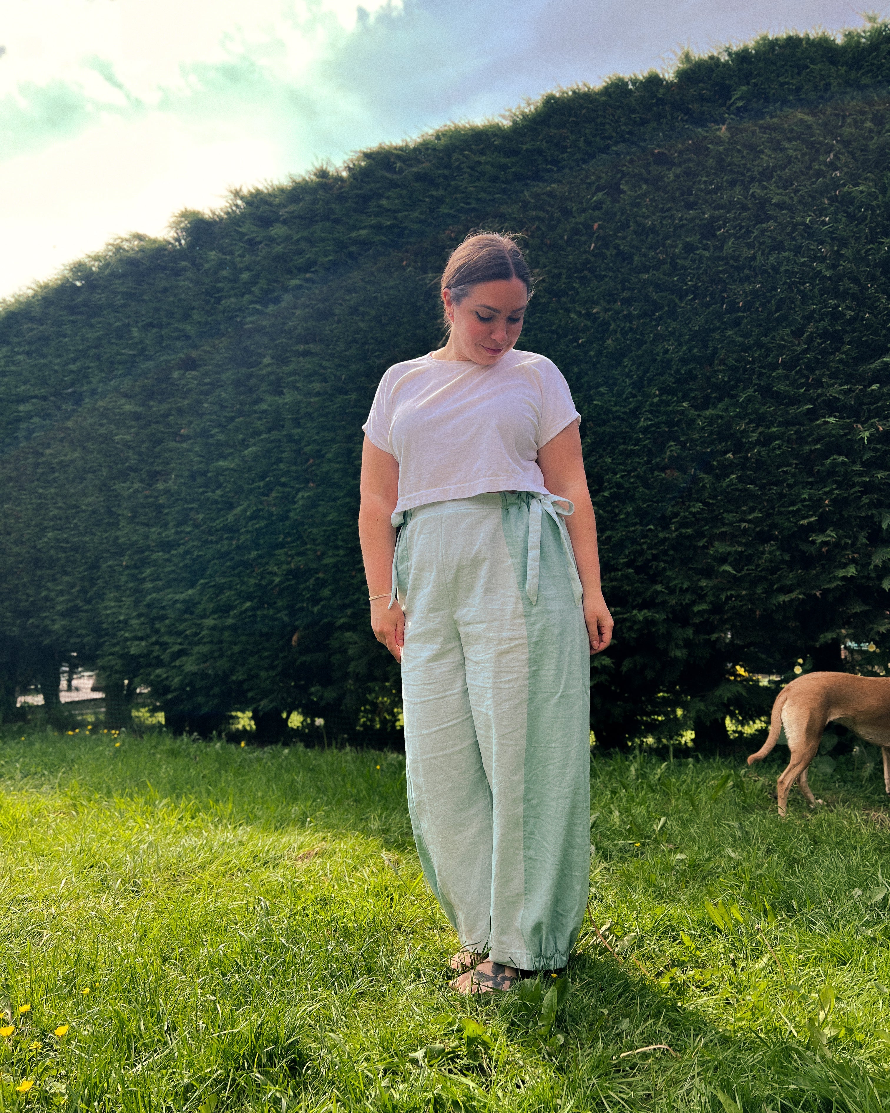 A woman standing on a grassy lawn in front of a large green hedge, wearing a white t-shirt and light sage green wide-leg pants with a tie waist. A dog is visible on the right side of the background.