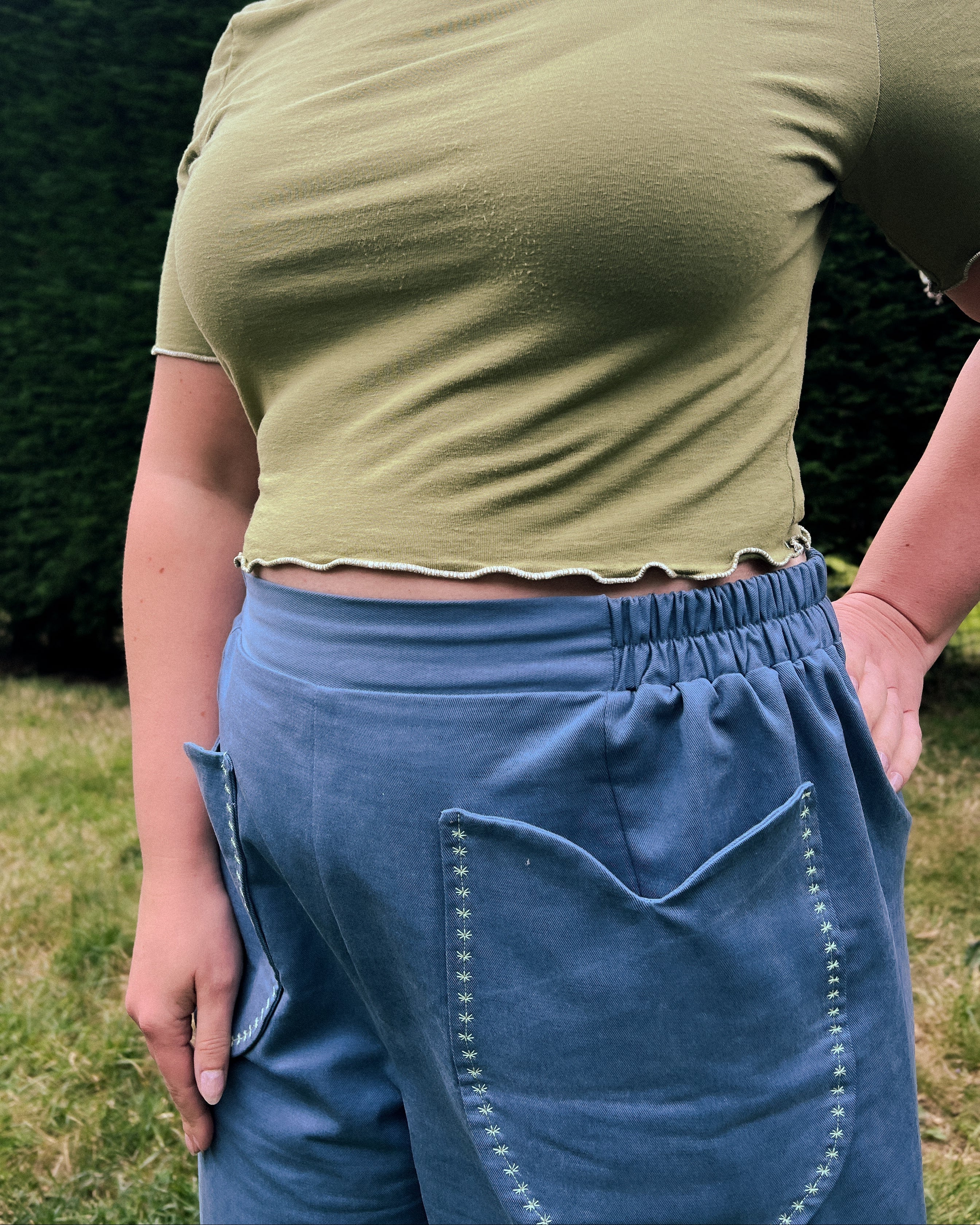 A person outdoors wearing a cropped olive green short-sleeve top and blue denim shorts, photographed from the back with a hedge and grass in the background.