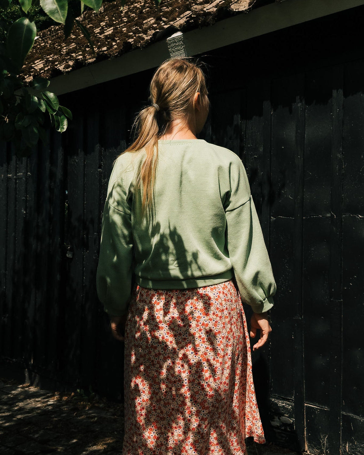 Person wearing a green sweater and patterned skirt standing in front of a dark wall.