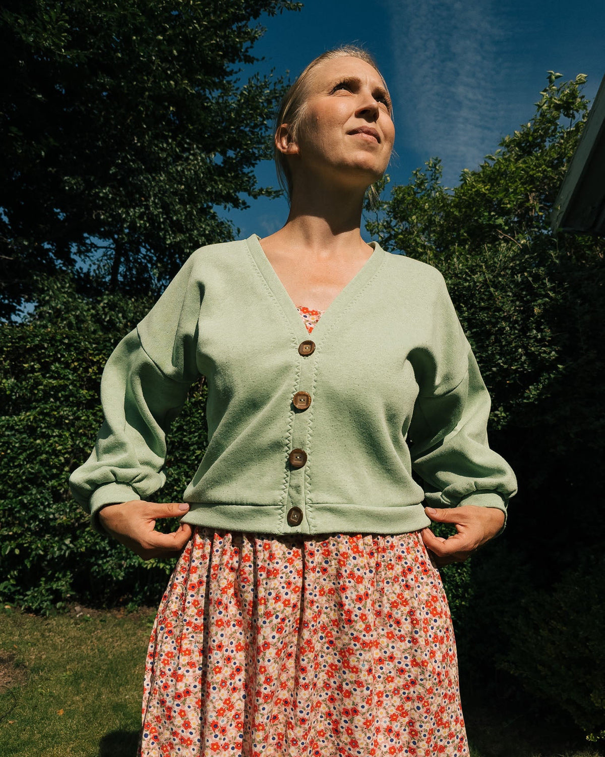 Woman wearing a light green cardigan and floral skirt outdoors with trees and blue sky in the background.