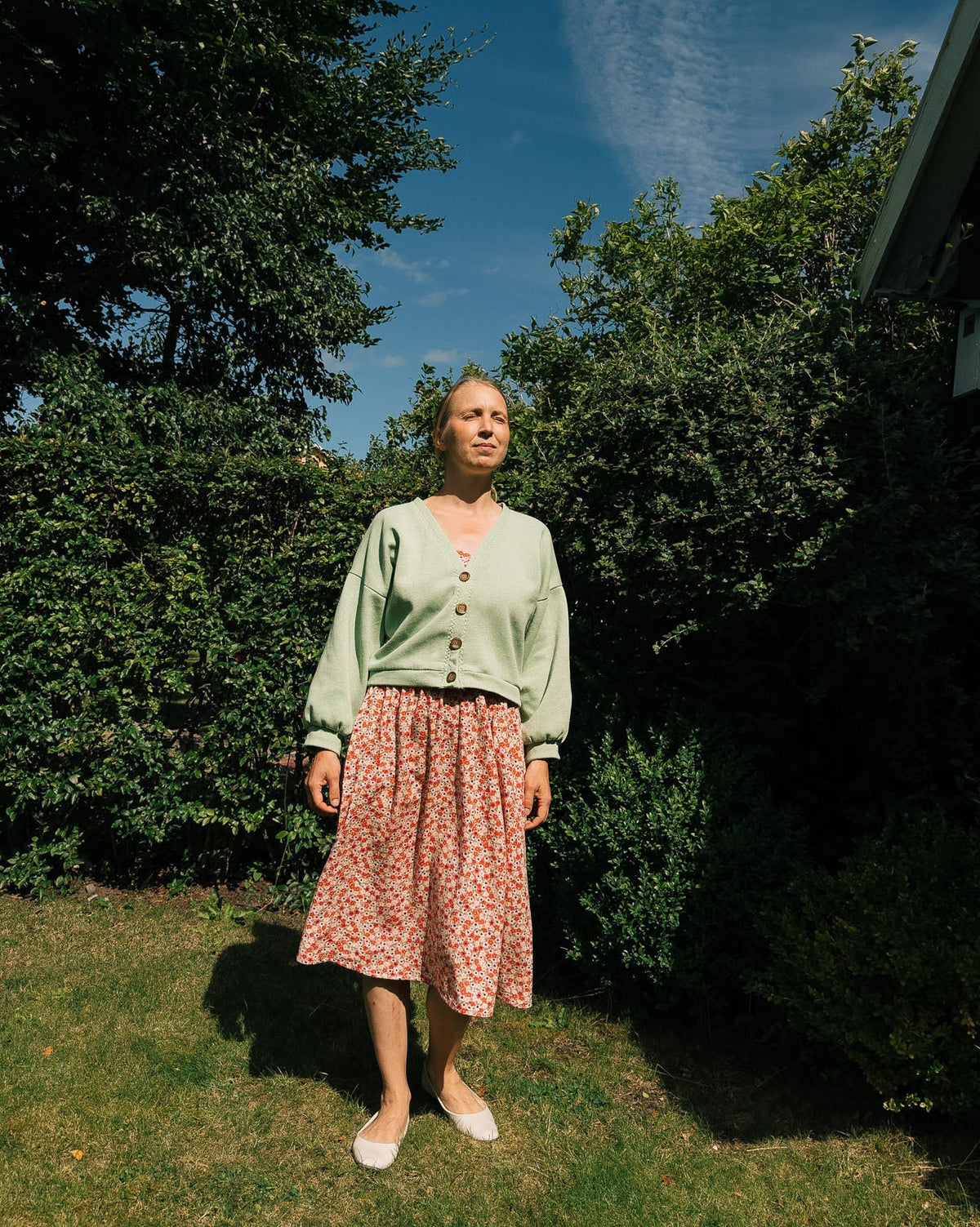 Woman standing outdoors in a garden with trees and a clear sky.