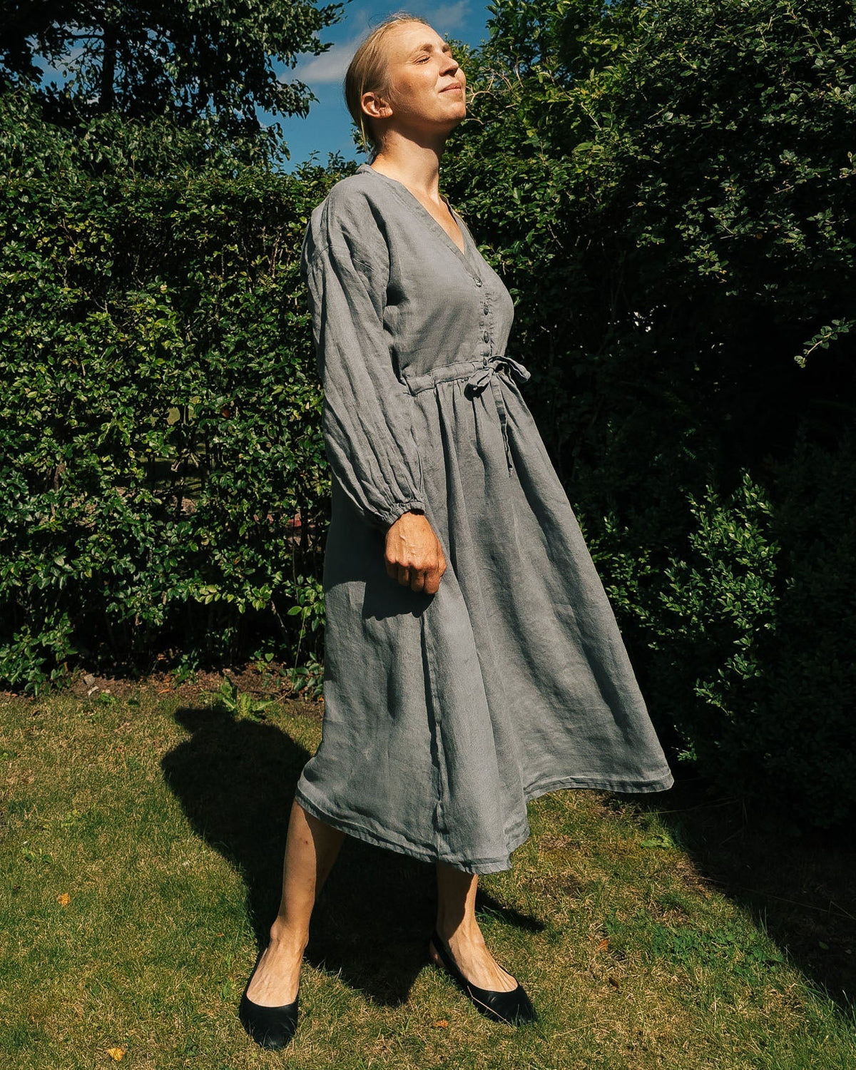 Woman in a gray dress standing outdoors with trees and blue sky in the background