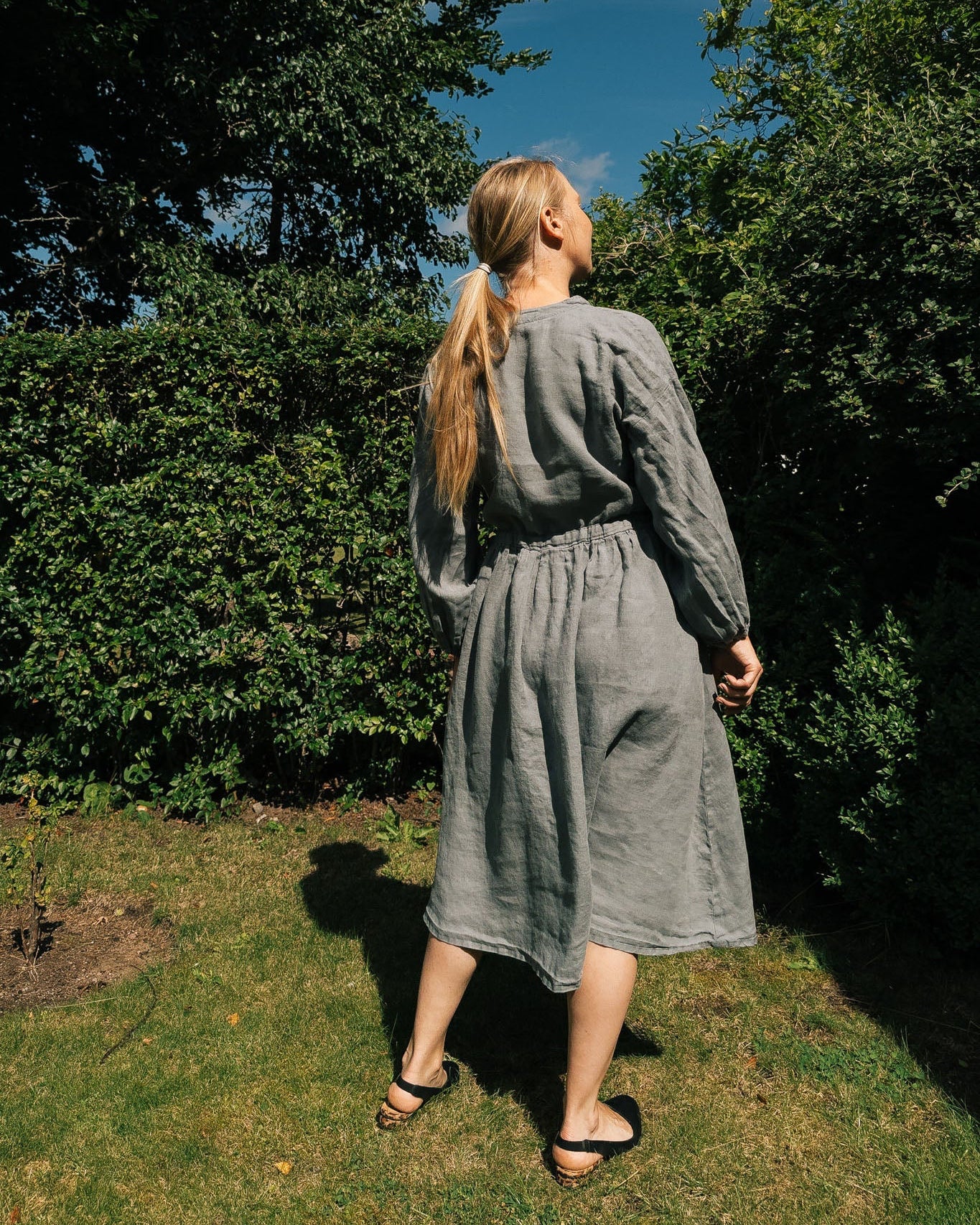 Woman in a gray dress standing in a garden with trees and blue sky.