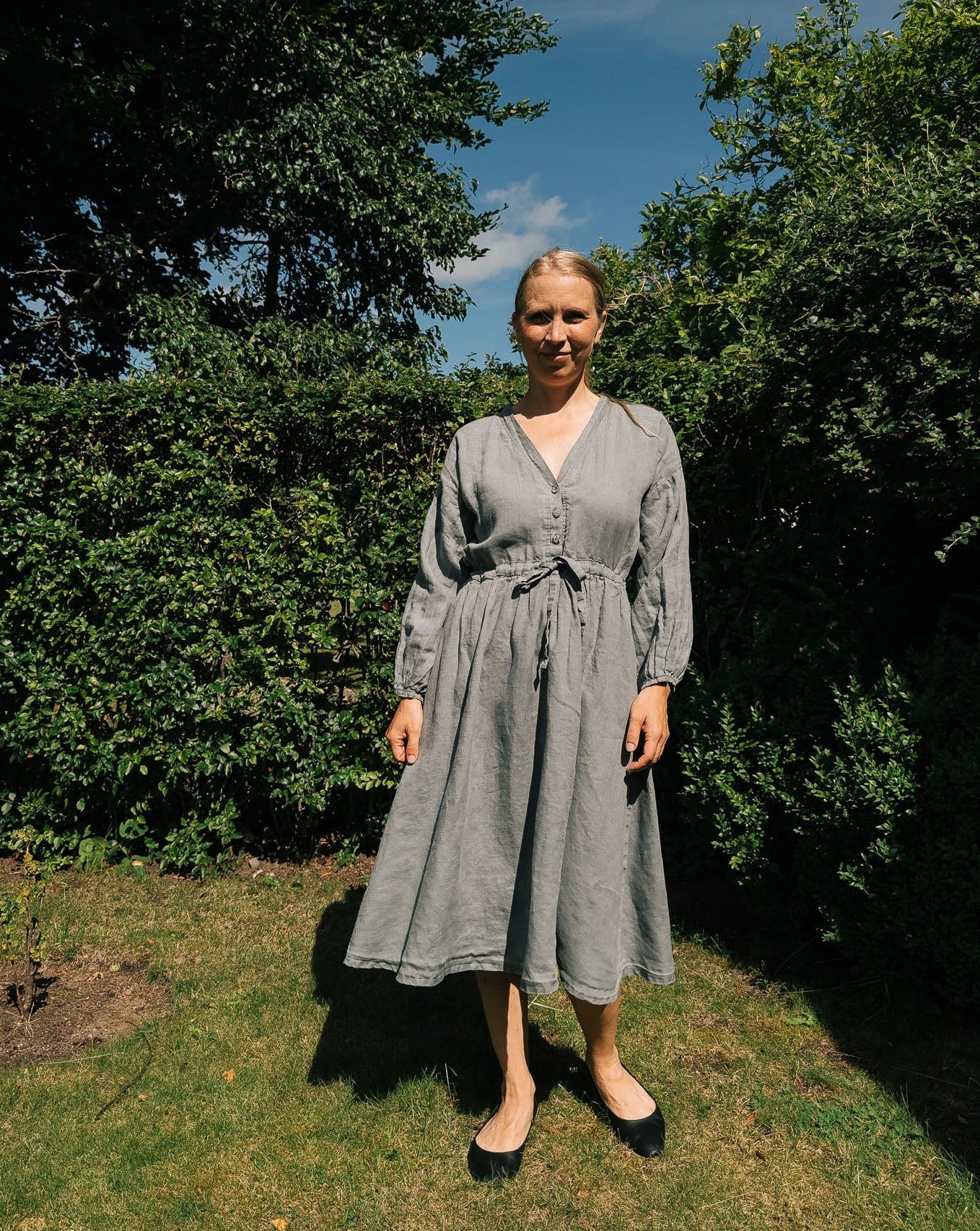 Woman in a gray dress standing outdoors with greenery and blue sky in the background
