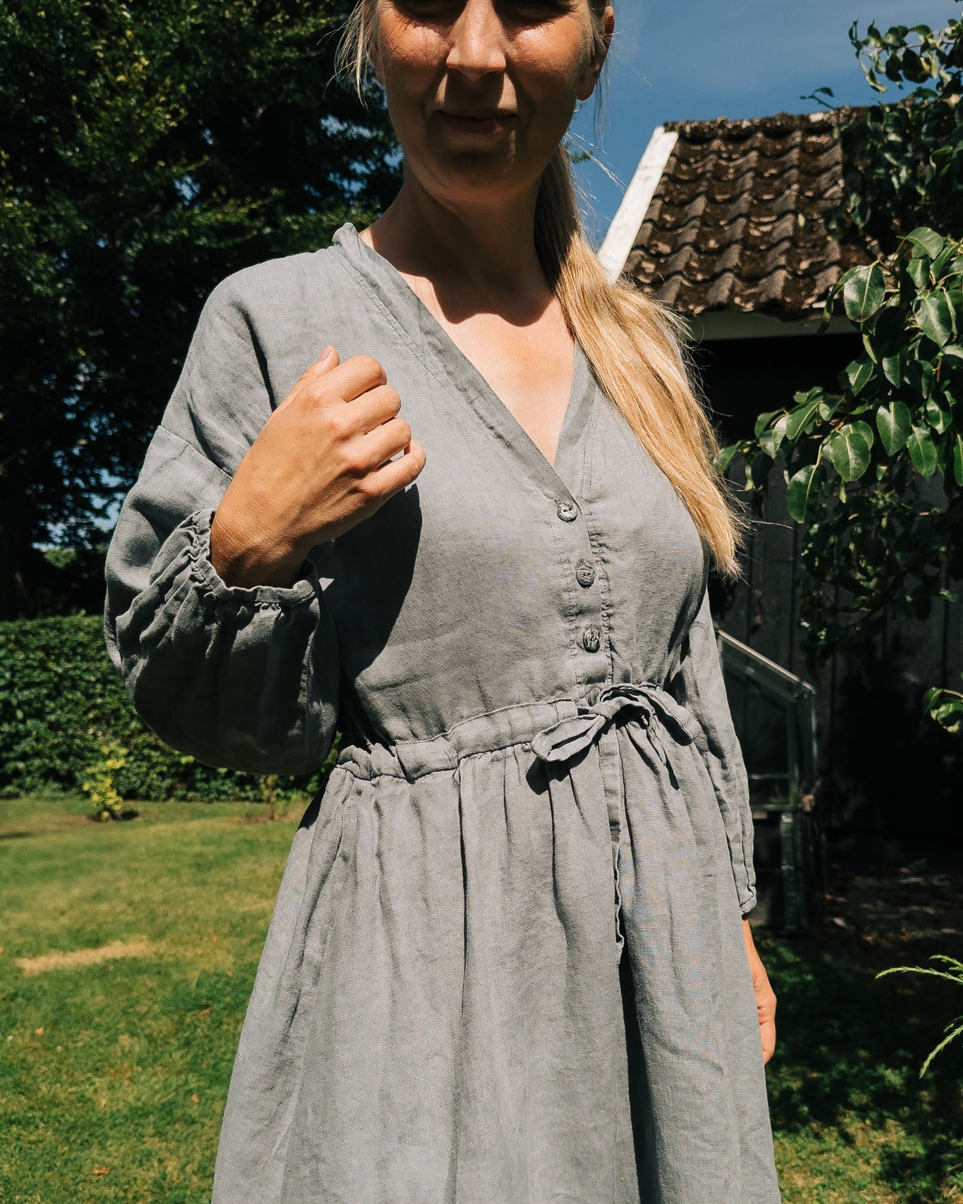 Woman wearing a gray dress standing outdoors with greenery and a building in the background