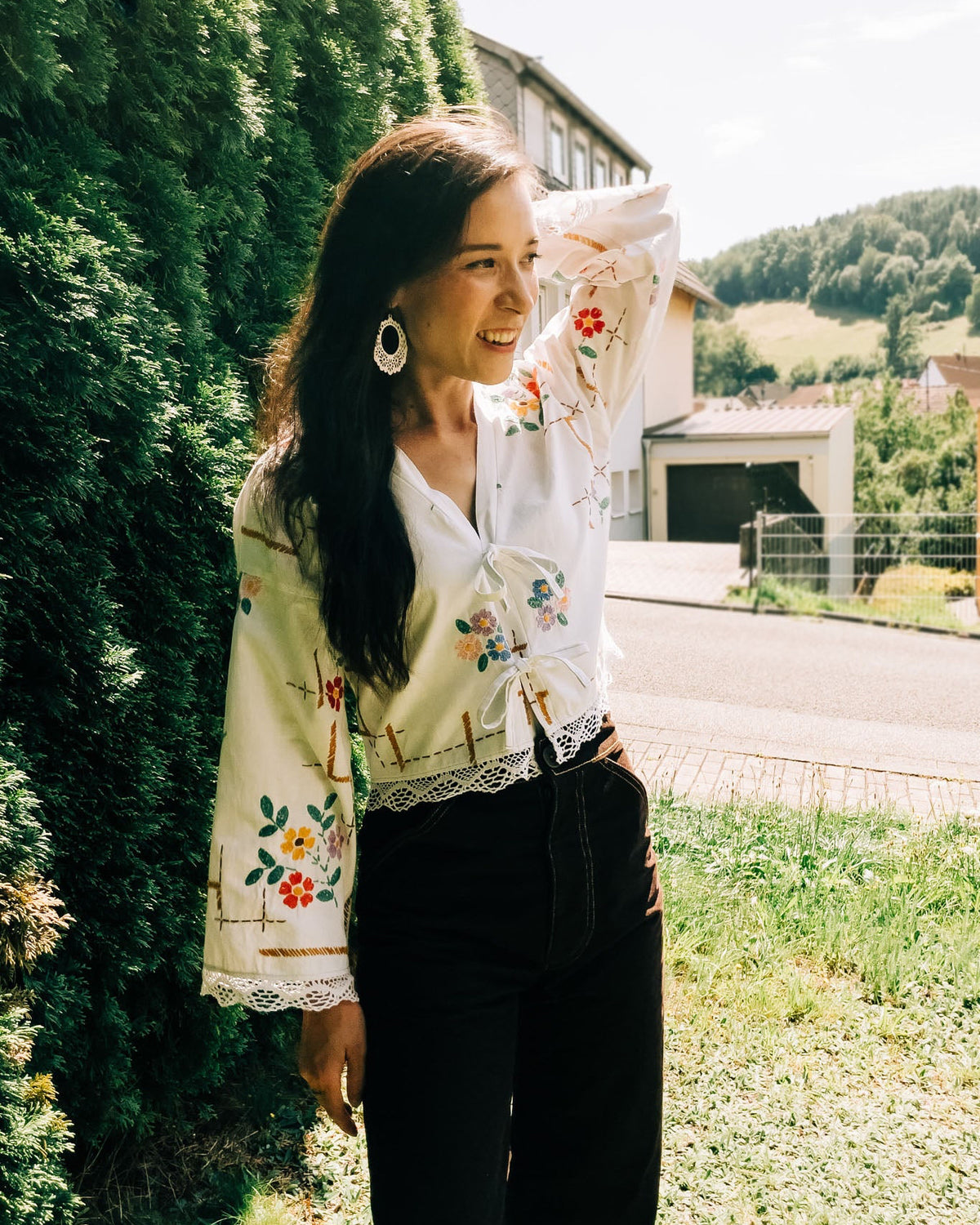 Woman wearing a traditional embroidered jacket outdoors with greenery and a house in the background