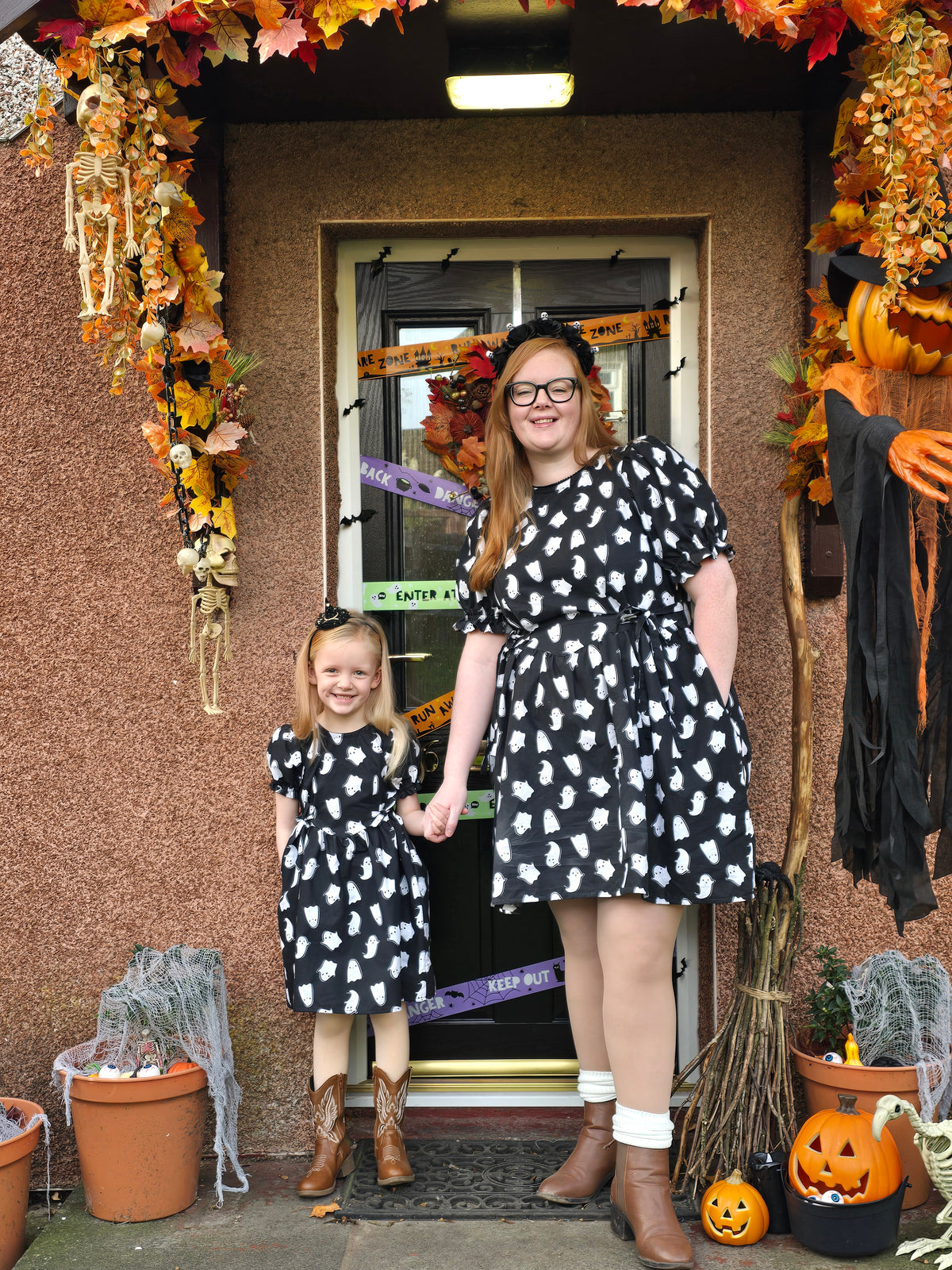 A woman and a young girl stand holding hands in front of a decorated front door, wearing matching black dresses with a white cat pattern. The scene is heavily styled for Halloween with autumn leaves, pumpkins, cobwebs, and a black broom.