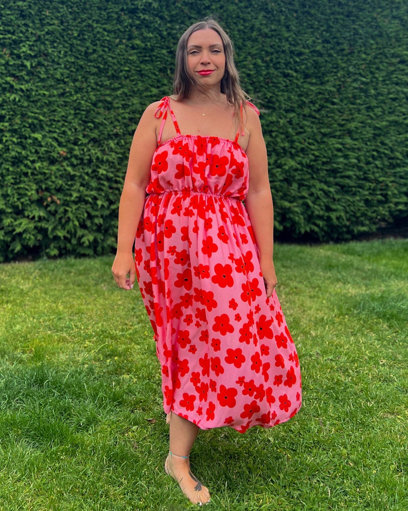 Woman in a red floral dress standing in front of a green hedge