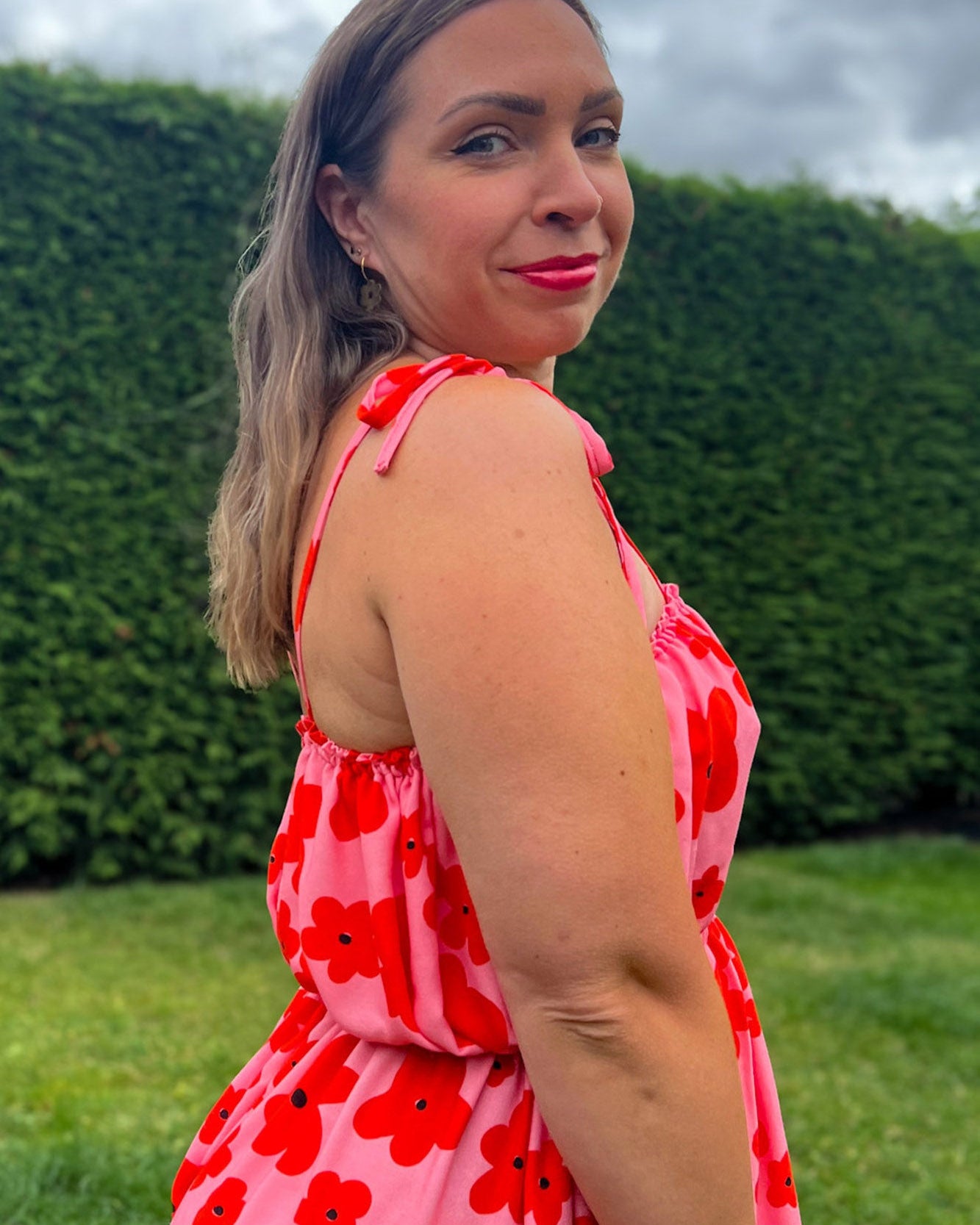 Woman wearing a red floral dress standing outdoors with green hedges and grass in the background
