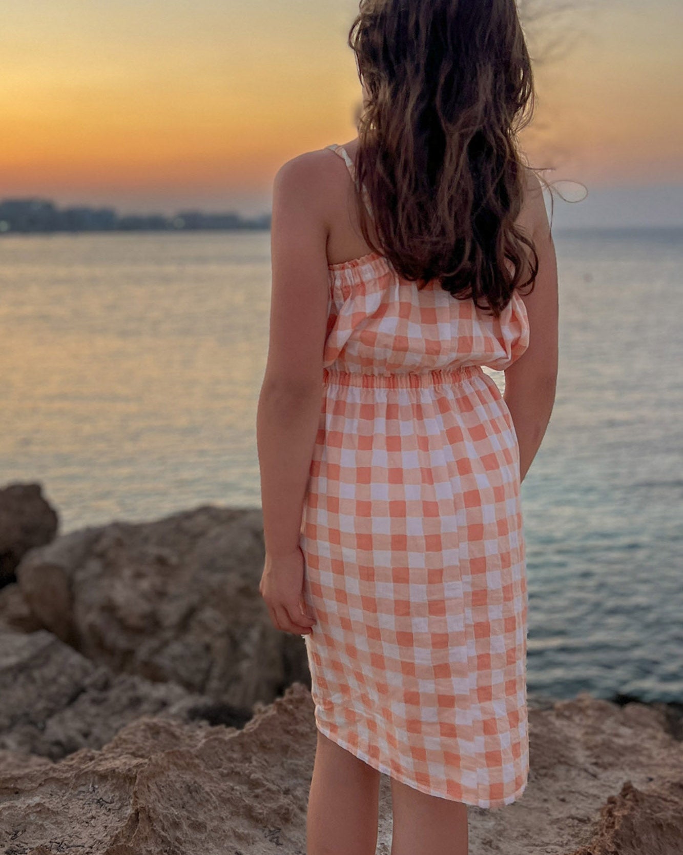 Child in a checkered dress standing on a rocky beach at sunset.