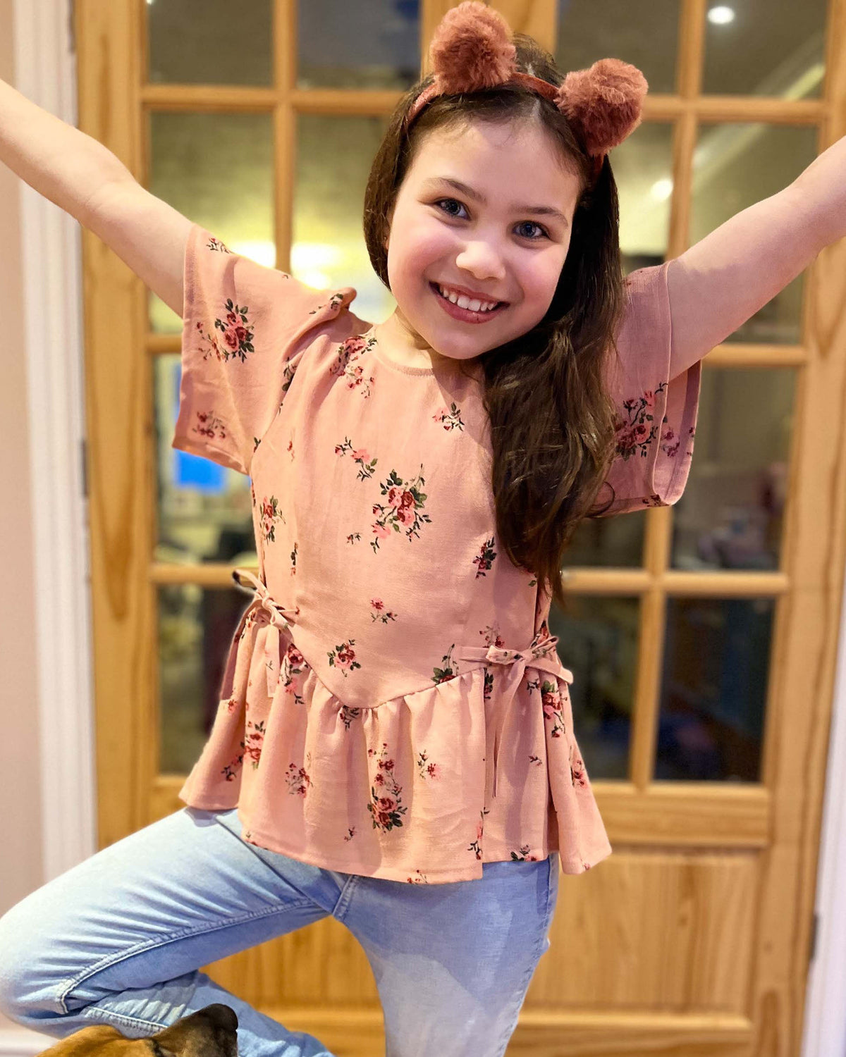 Child in a pink floral top and blue jeans with animal ears, standing indoors.