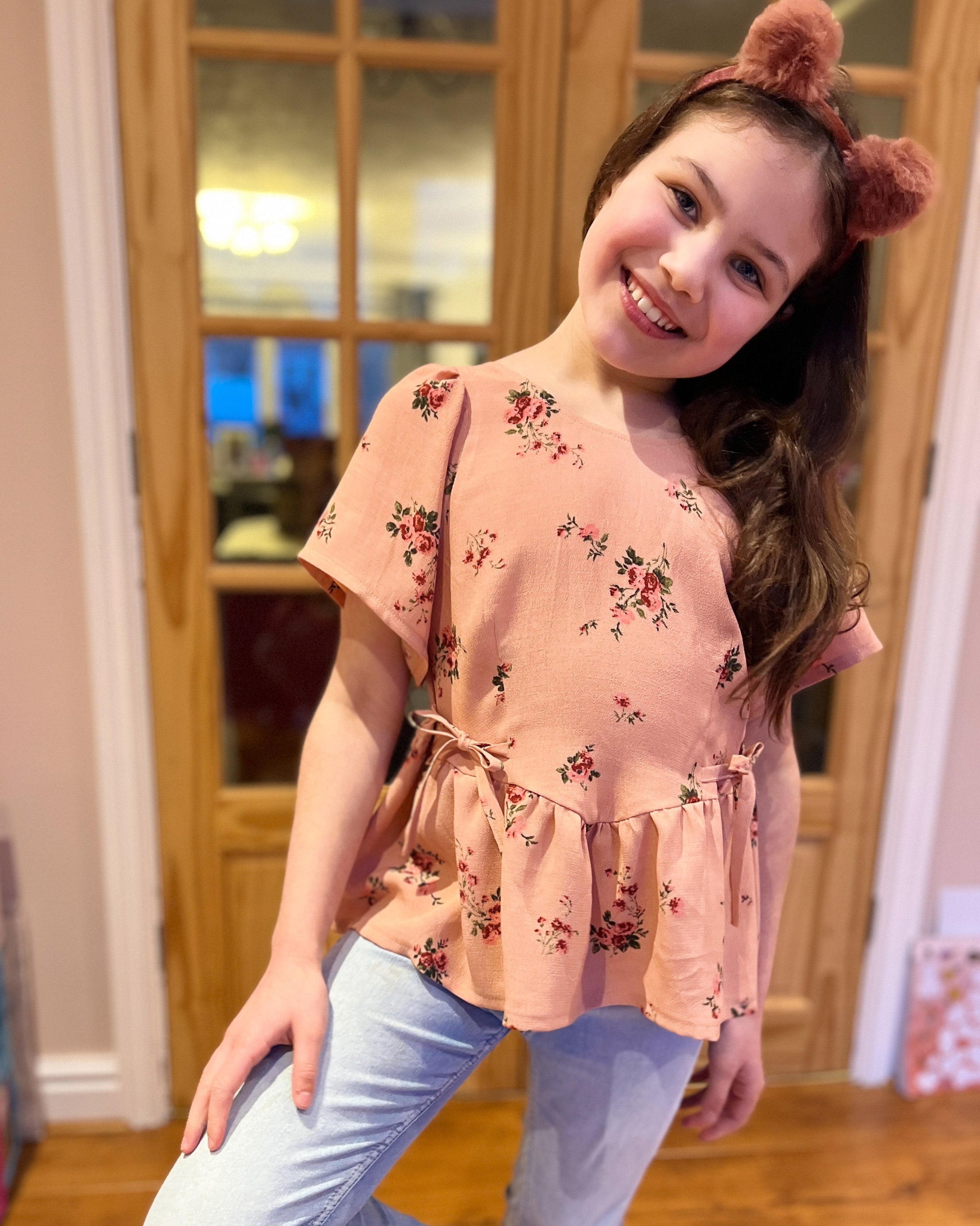 Young girl in a pink floral top and blue jeans standing indoors.