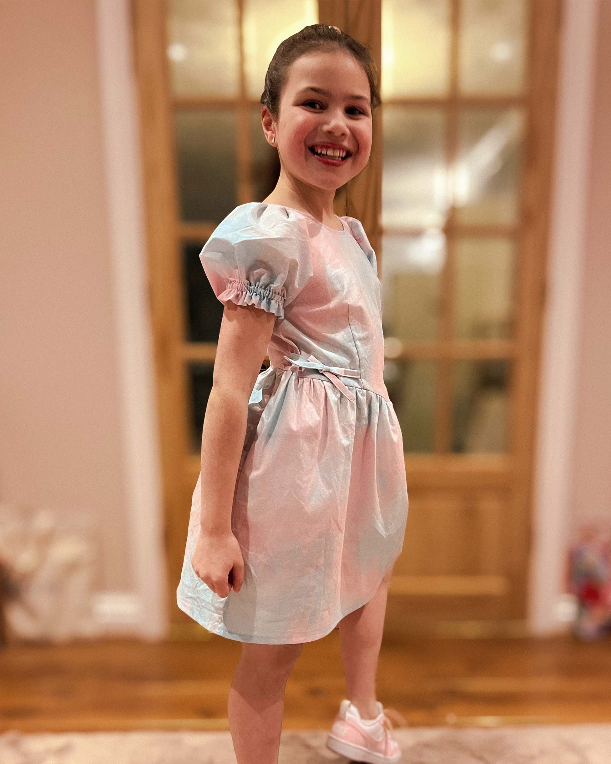 Young girl in a pink and white dress standing in a room with wooden flooring and a large mirror.