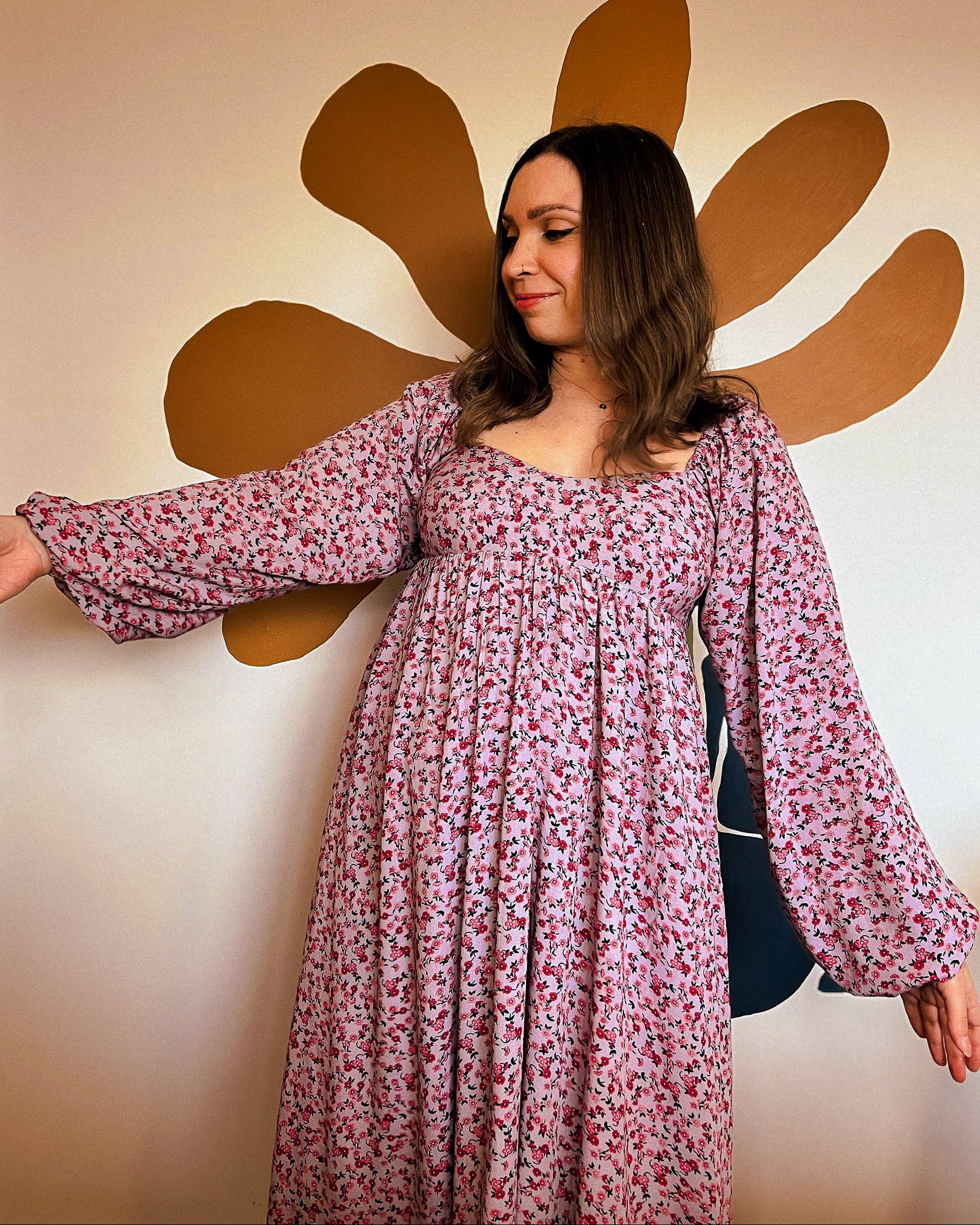 Woman in a floral dress standing next to a large brown flower on a beige wall.