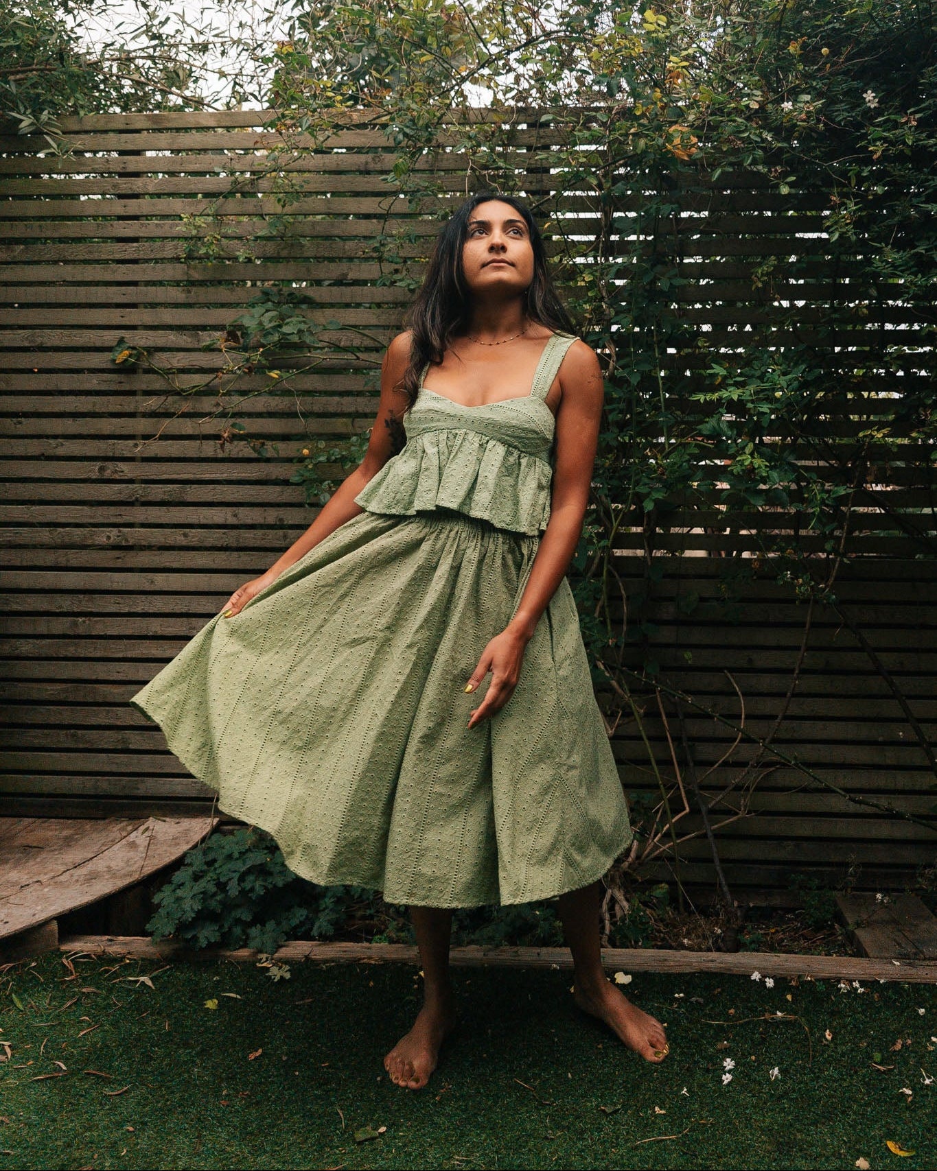Woman wearing a green dress standing outdoors with a wooden fence and trees in the background