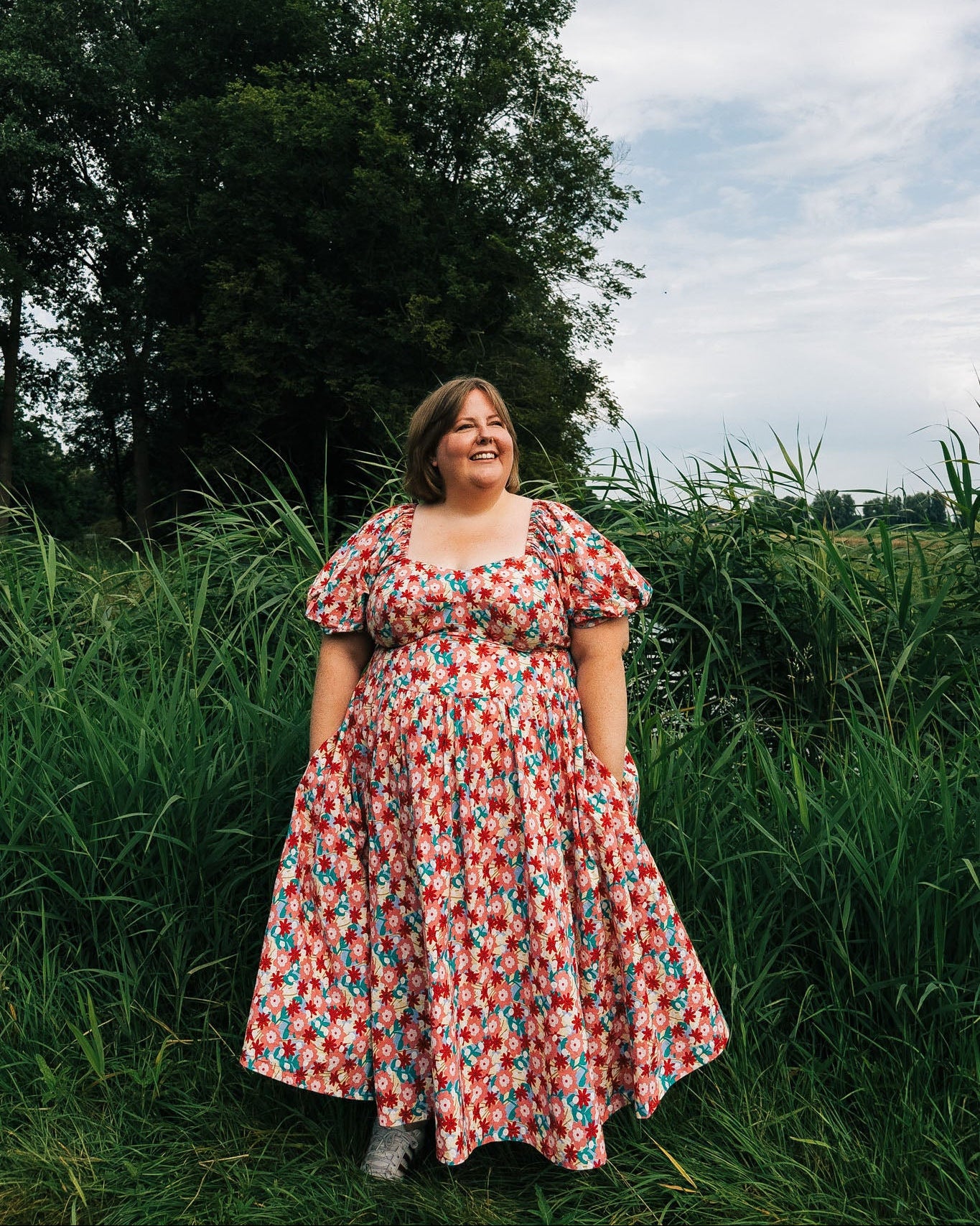 Woman in a floral dress standing in a grassy field with trees and blue sky in the background