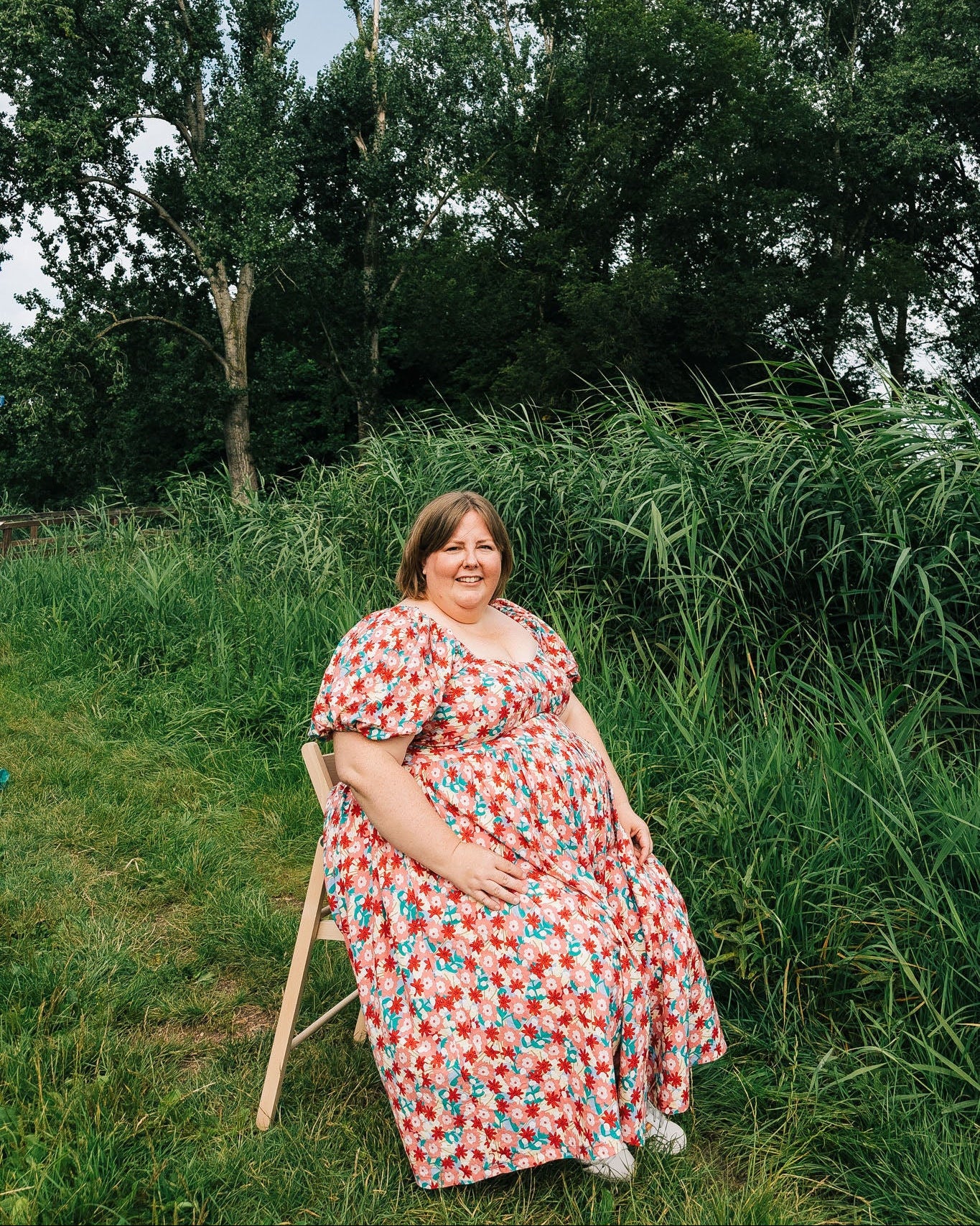 Woman in a floral dress sitting on a chair in a grassy field with trees in the background