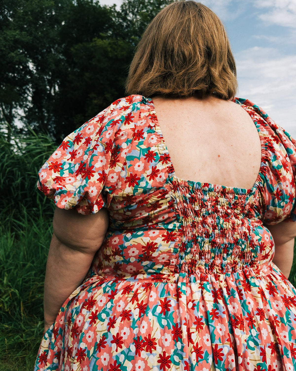 Person wearing a colorful floral dress standing outdoors with trees and sky in the background
