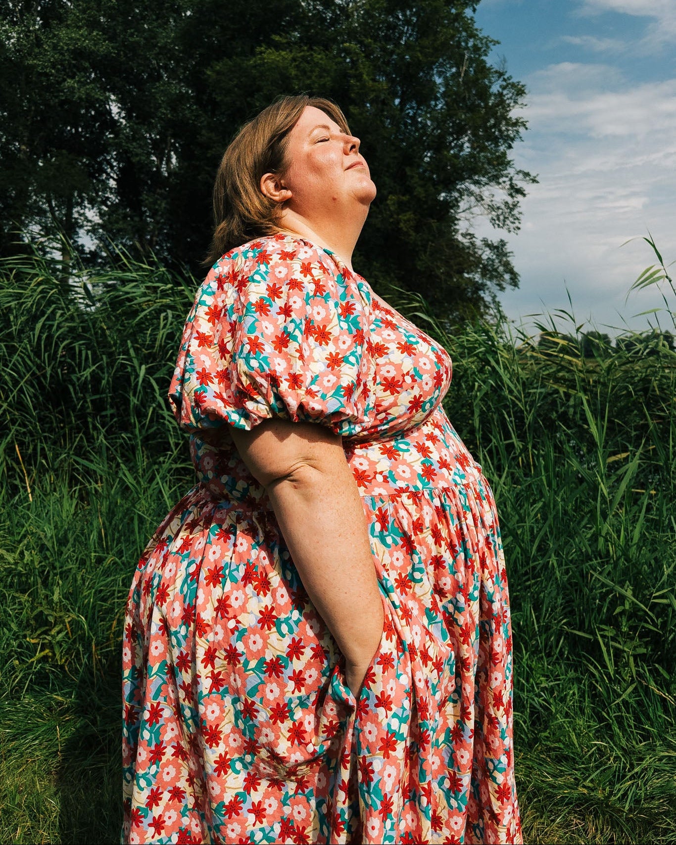 Woman in a floral dress standing in a grassy field with trees and blue sky in the background