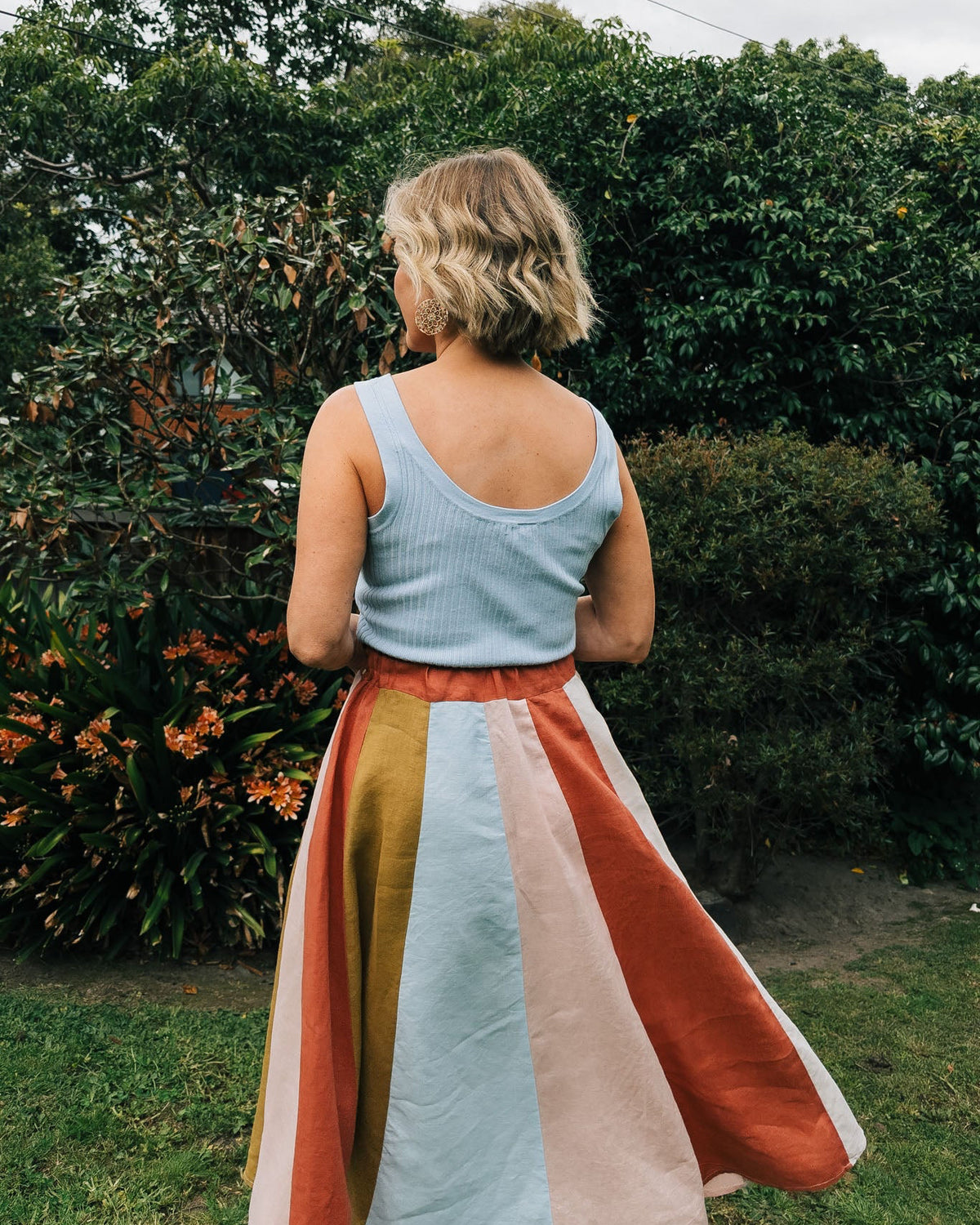 Woman standing outdoors with a colorful skirt and light blue top, surrounded by greenery.