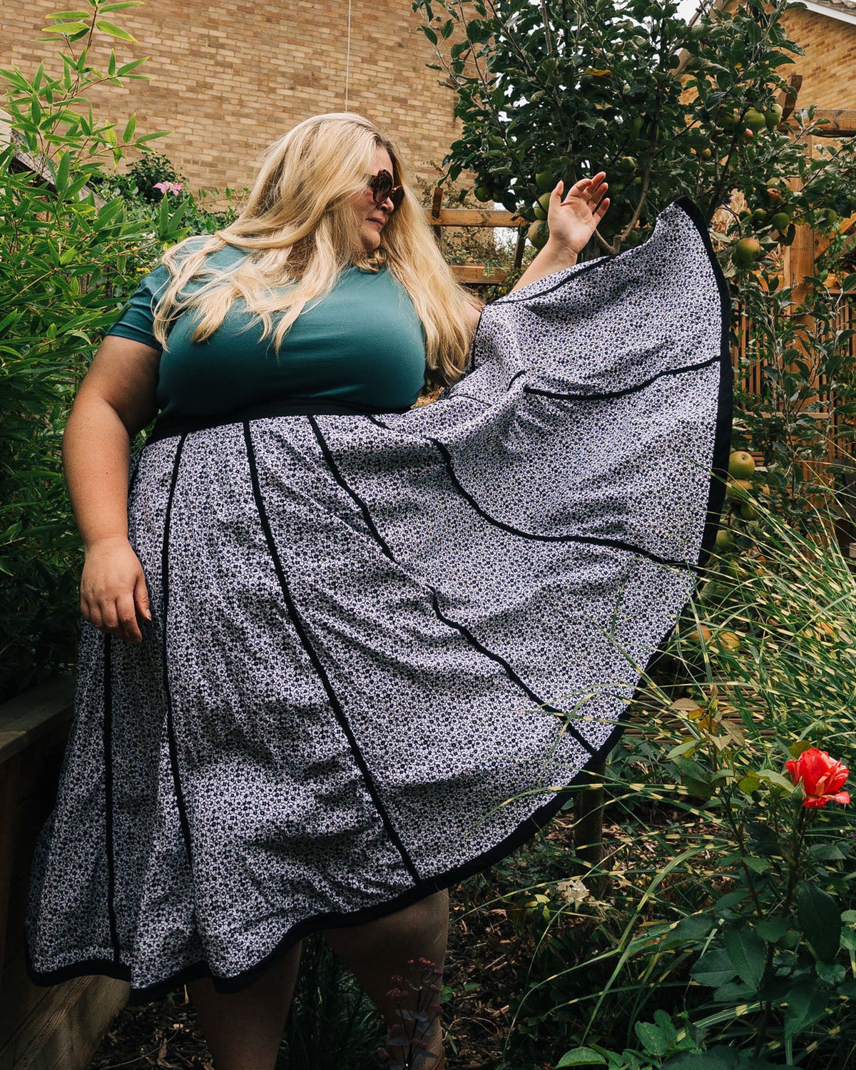 Woman wearing a teal top and black and white patterned skirt in a garden setting
