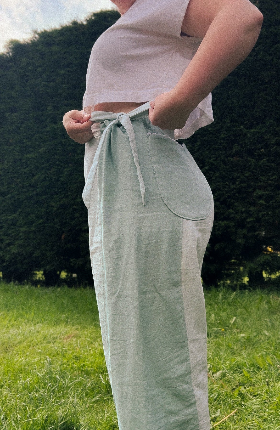 A person outdoors wearing light sage green linen wide-leg pants with a tie waist and a white cropped tank top, standing on a grassy lawn with trees in the background.