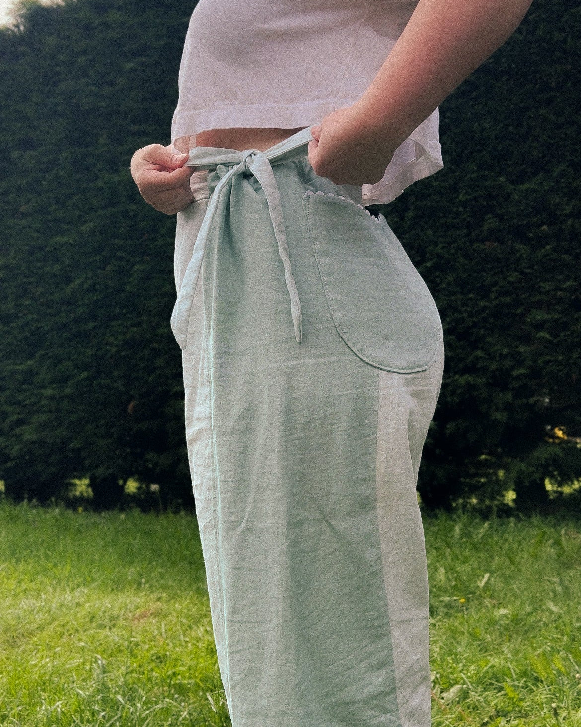 A person outdoors wearing light sage green linen wide-leg pants with a tie waist and a white cropped tank top, standing on a grassy lawn with trees in the background.
