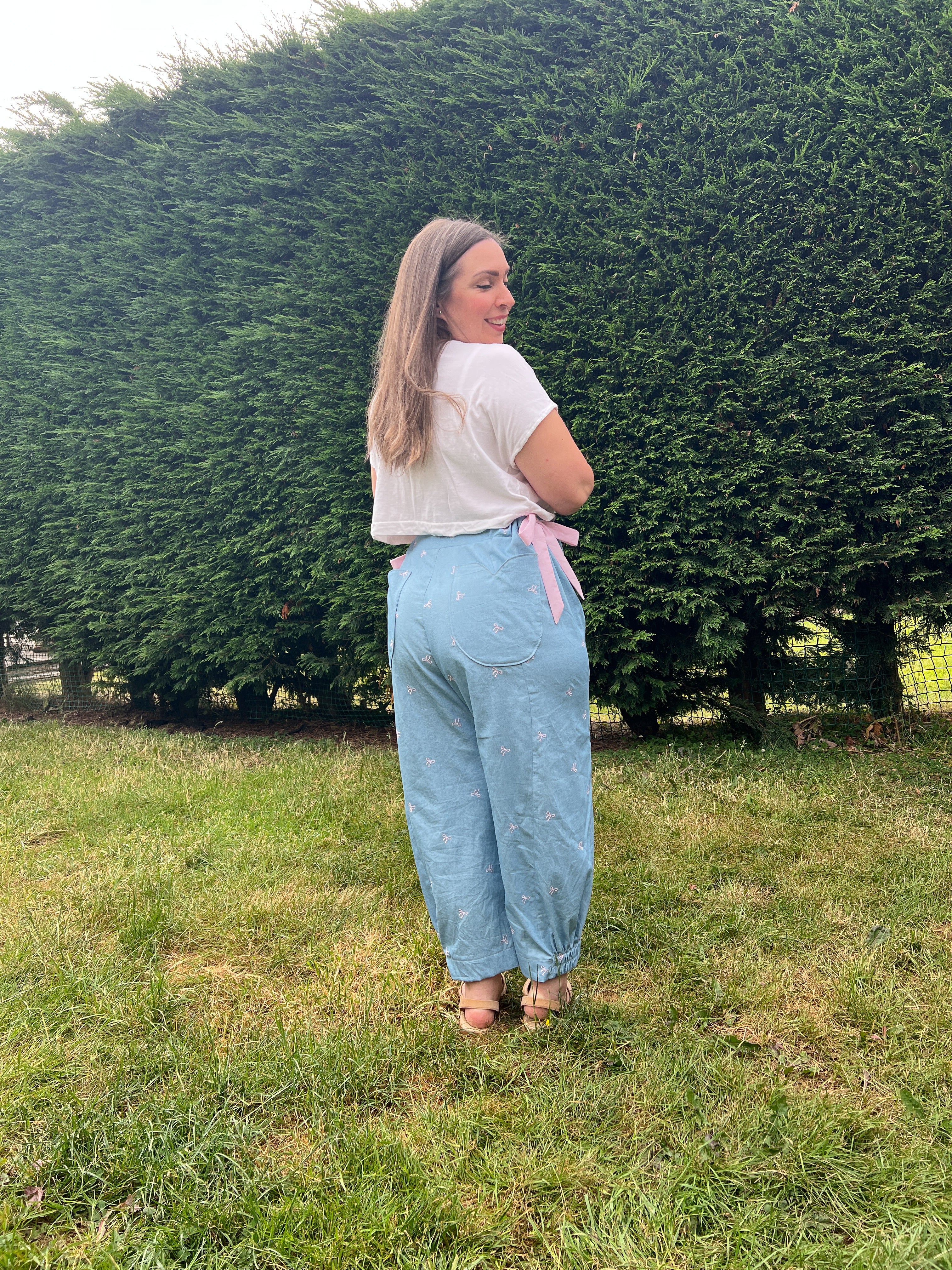 A woman stands on a grassy lawn in front of a large green hedge, wearing light blue wide-leg jeans with a pink ribbon tie at the waist, a white t-shirt, and brown sandals.
