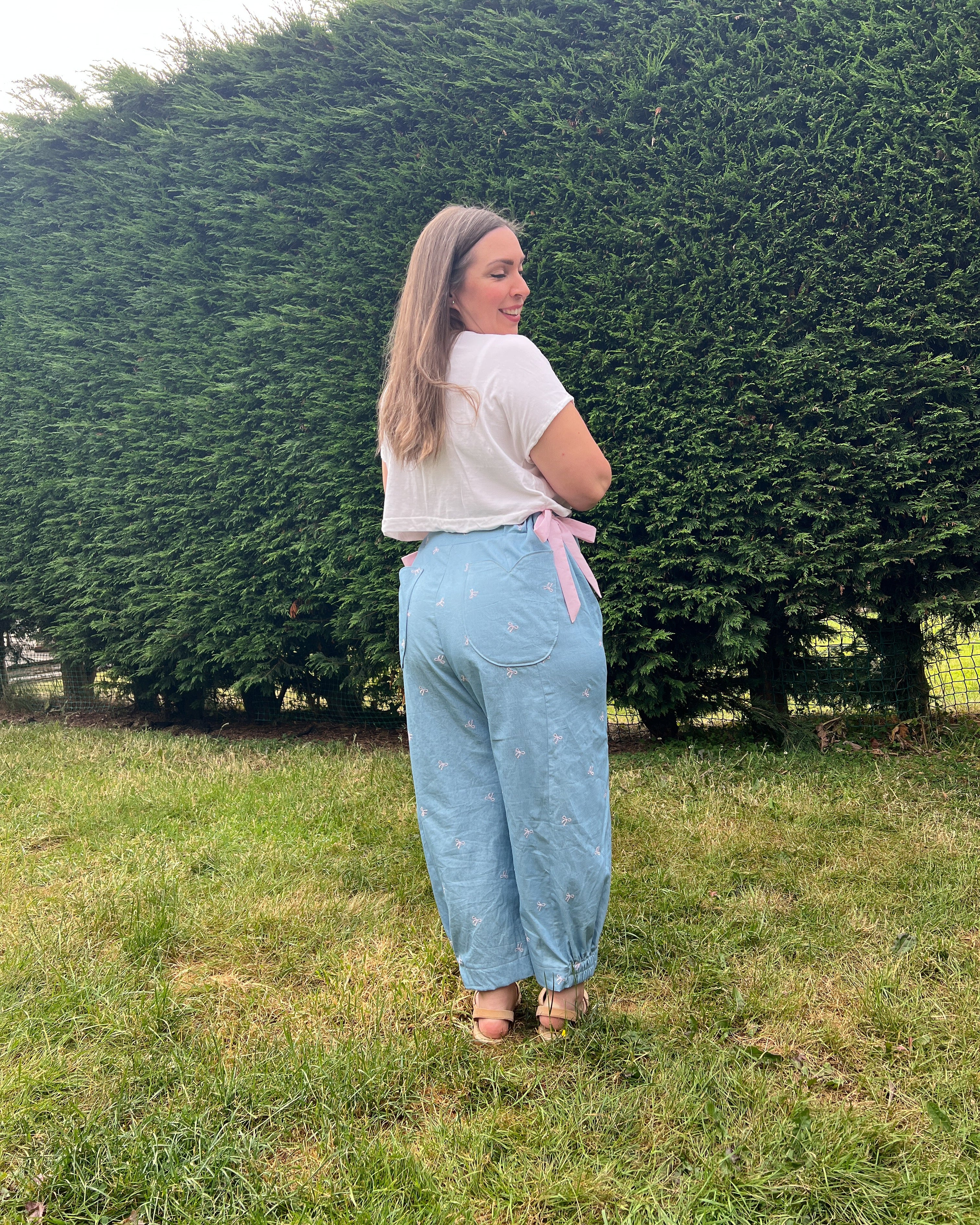 A woman stands on a grassy lawn in front of a large green hedge, wearing light blue wide-leg jeans with a pink ribbon tie at the waist, a white t-shirt, and brown sandals.