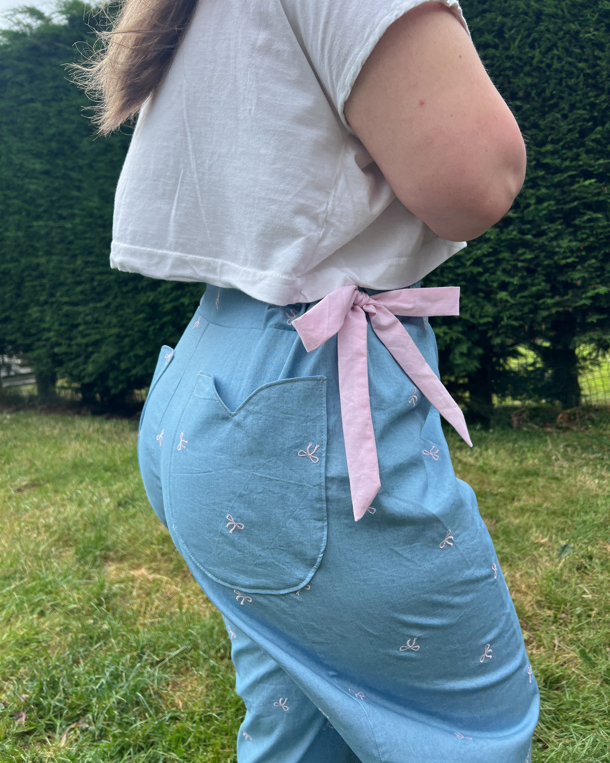 A person outdoors wearing light blue embroidered pants with a pink ribbon tied at the back waist, paired with a white cropped T-shirt.