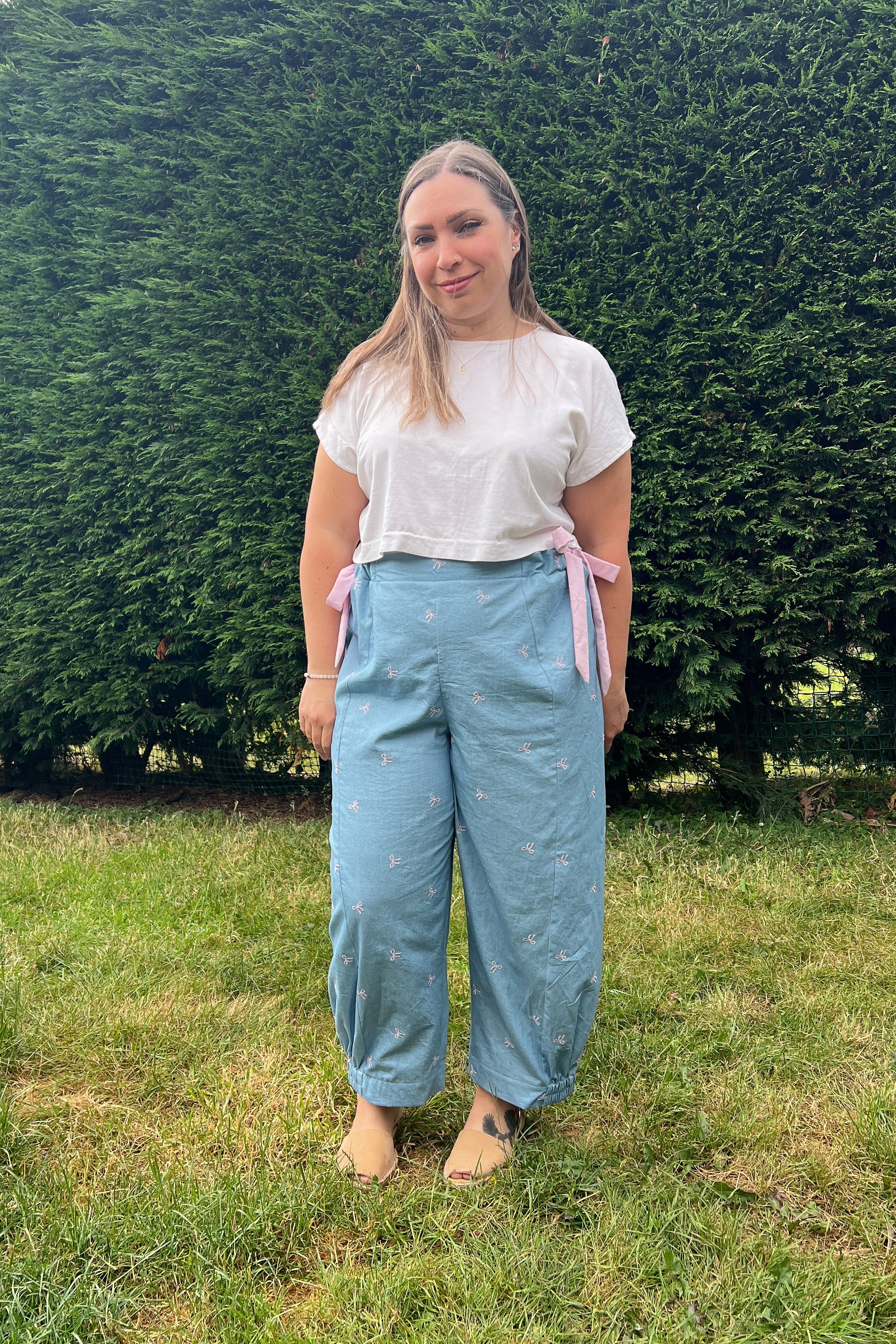 A woman standing outdoors on a grassy lawn in front of a large green hedge, wearing light blue wide-leg pants with pink side ties, a white short-sleeve top, and bare feet.