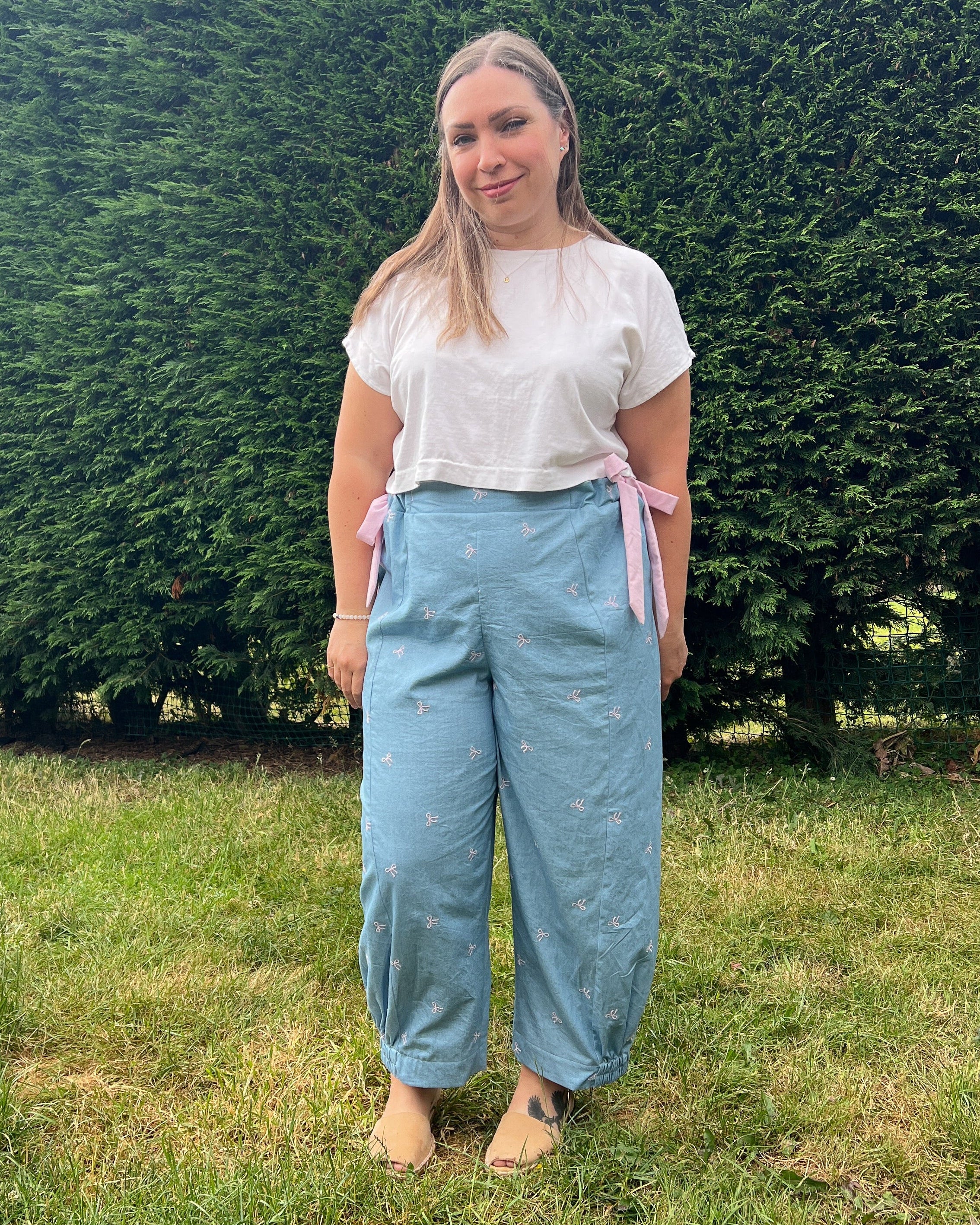 A woman standing outdoors on a grassy lawn in front of a large green hedge, wearing light blue wide-leg pants with pink side ties, a white short-sleeve top, and bare feet.