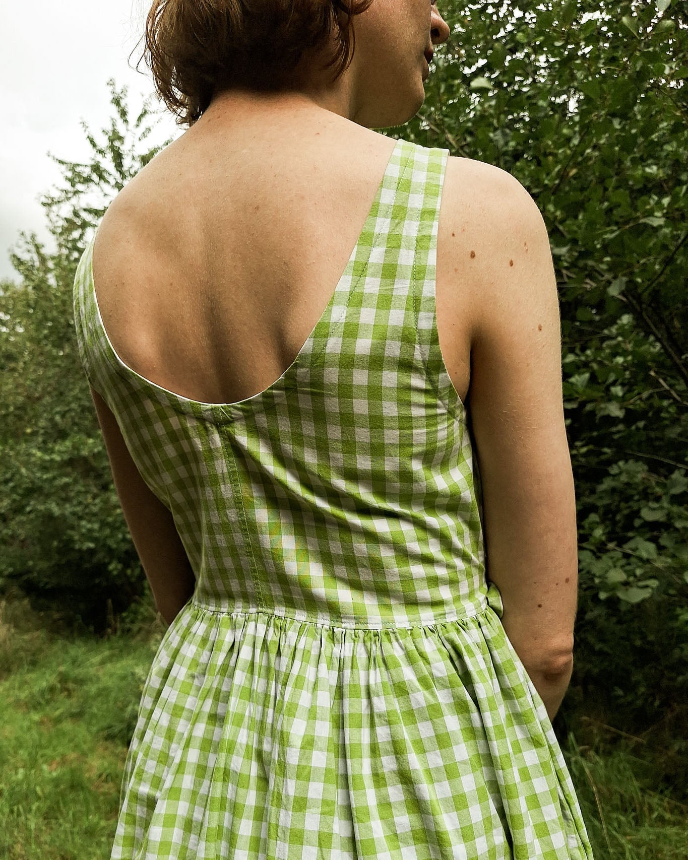 Person wearing a green checkered dress standing in a natural setting with trees and grass.