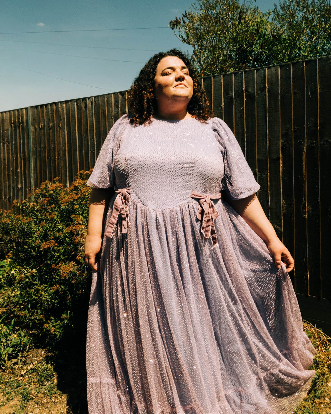 Woman in a long, light purple dress standing outdoors with a wooden fence and trees in the background.