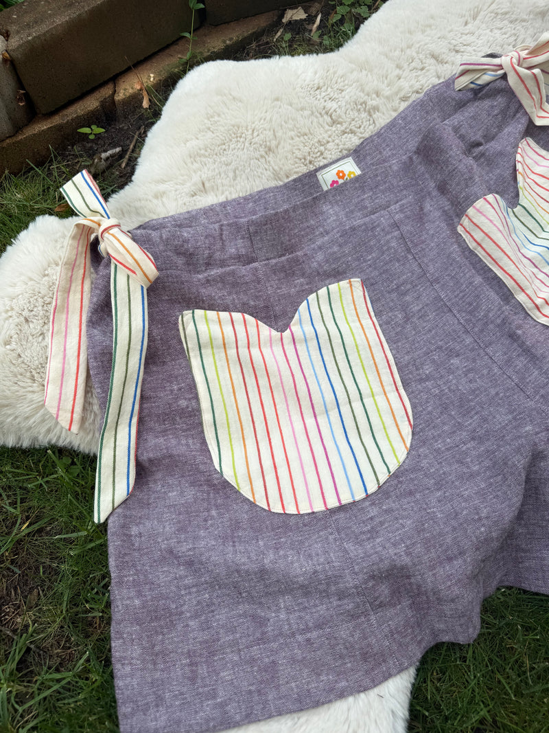 A lavender linen shorts with colorful striped pockets and tie straps, laid flat on a white faux fur rug over green grass.