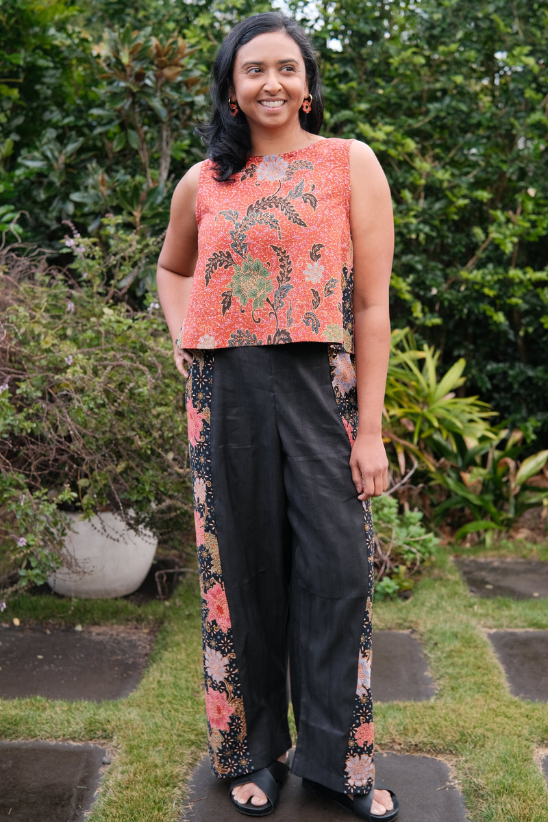 A woman stands outdoors in a garden wearing a sleeveless coral-orange top with a floral print and black wide-leg pants with matching floral side panels.