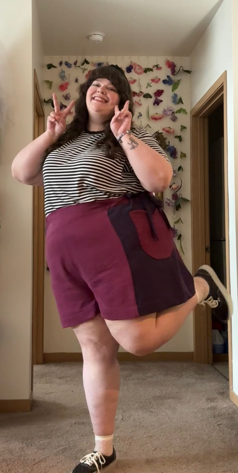 A woman posing in a home hallway wearing a black and white striped t-shirt and maroon colorblock shorts with a side pocket.