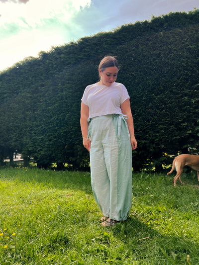 A woman standing on a grassy lawn in front of a large green hedge, wearing a white t-shirt and light sage green wide-leg pants with a tie waist. A dog is visible on the right side of the background.