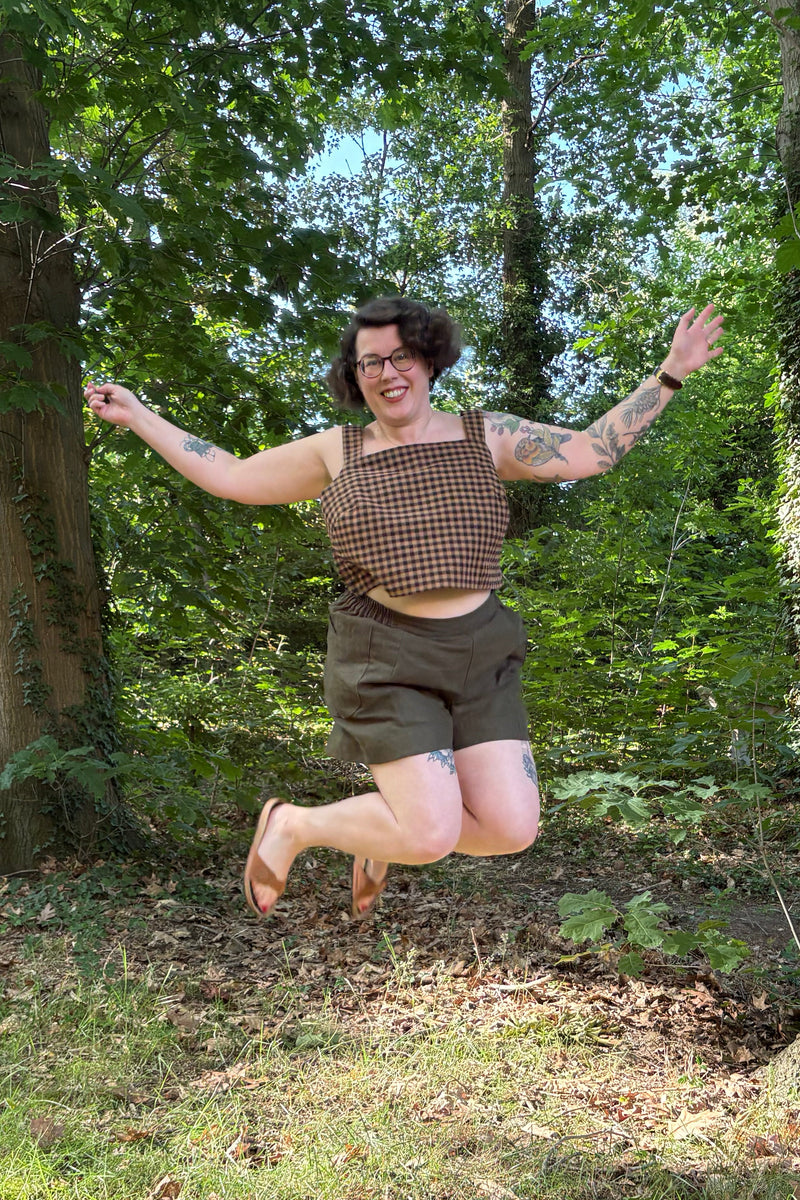 A woman wearing a brown checkered crop top and dark brown shorts jumps excitedly in a wooded outdoor setting with trees and grass visible.