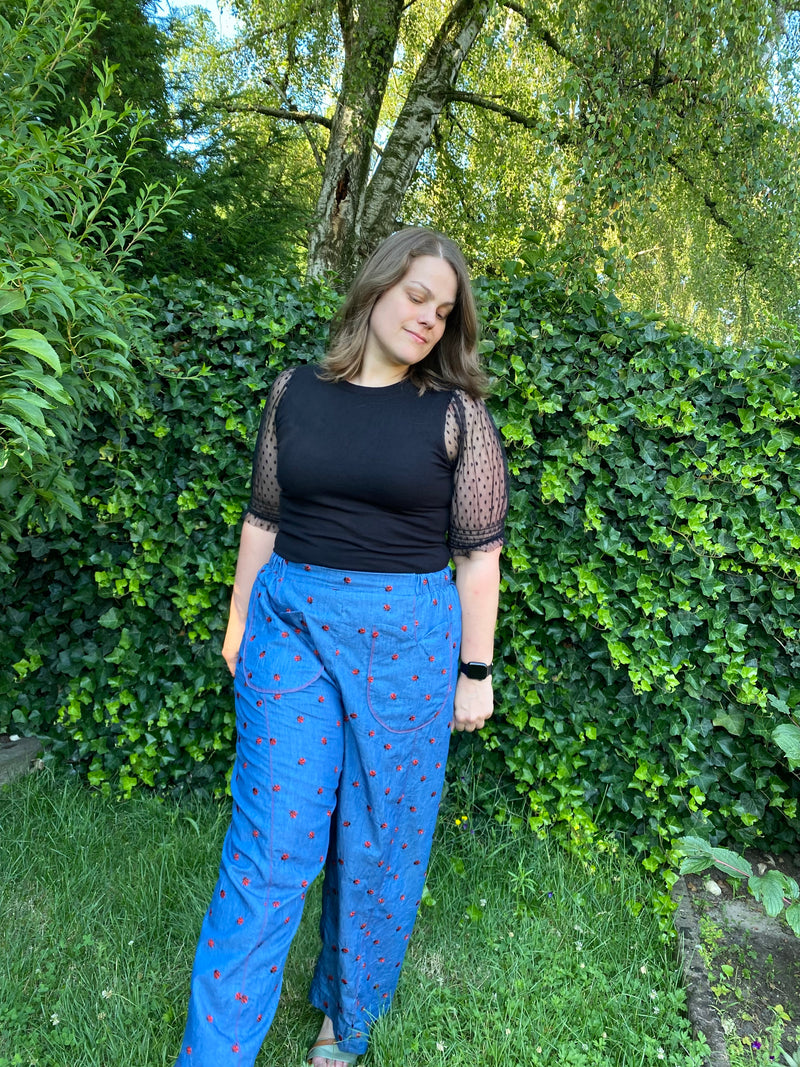 A woman standing outdoors in a garden wearing a black short-sleeve top with sheer patterned sleeves and blue wide-leg pants with a small red floral print.