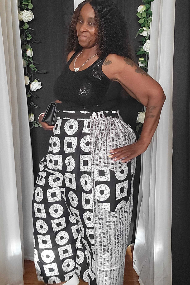 A woman posing in a black sleeveless top and high-waisted black-and-white patterned wide-leg pants, standing in front of a black curtain backdrop decorated with white flowers.