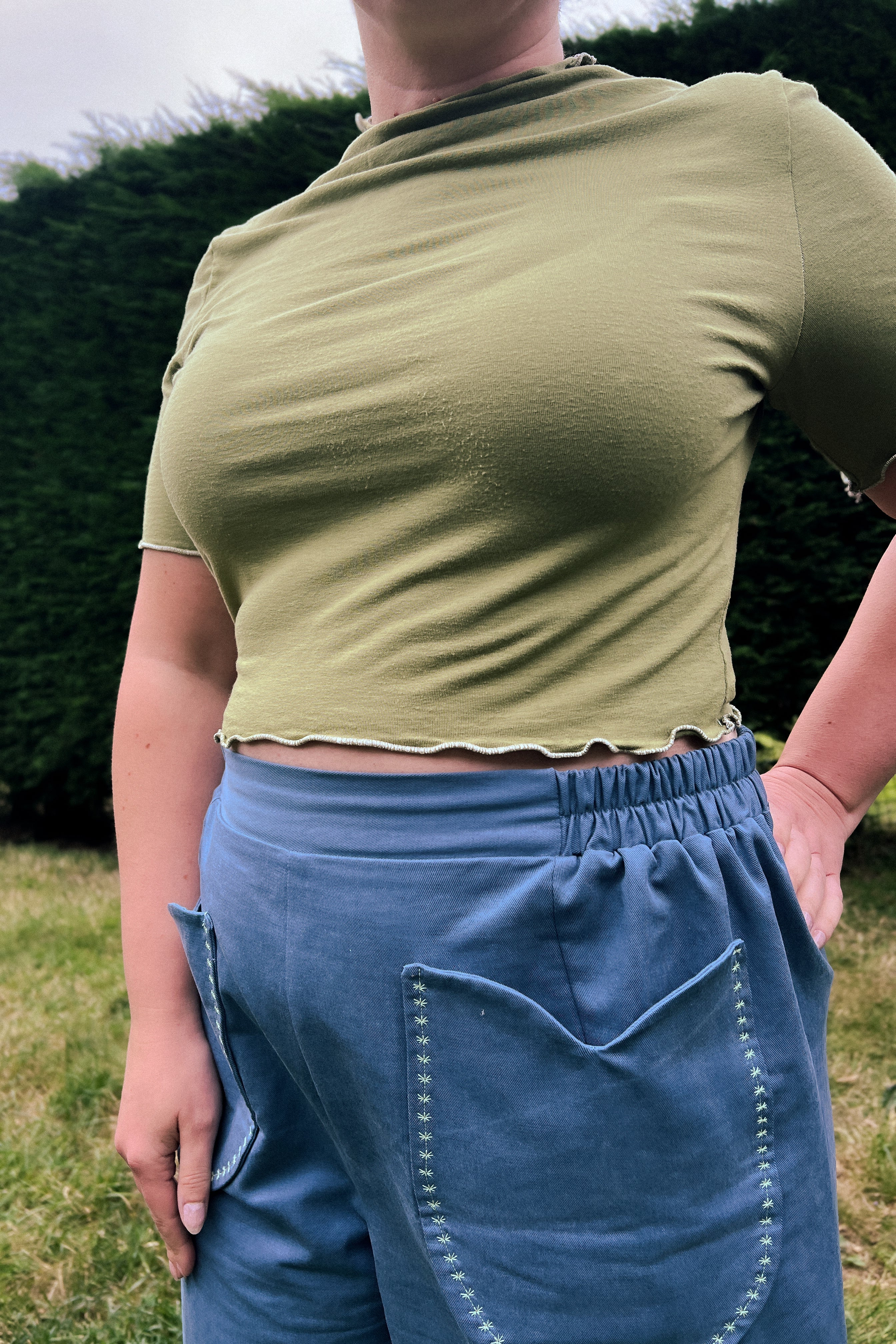A person outdoors wearing a cropped olive green short-sleeve top and blue denim shorts, photographed from the back with a hedge and grass in the background.