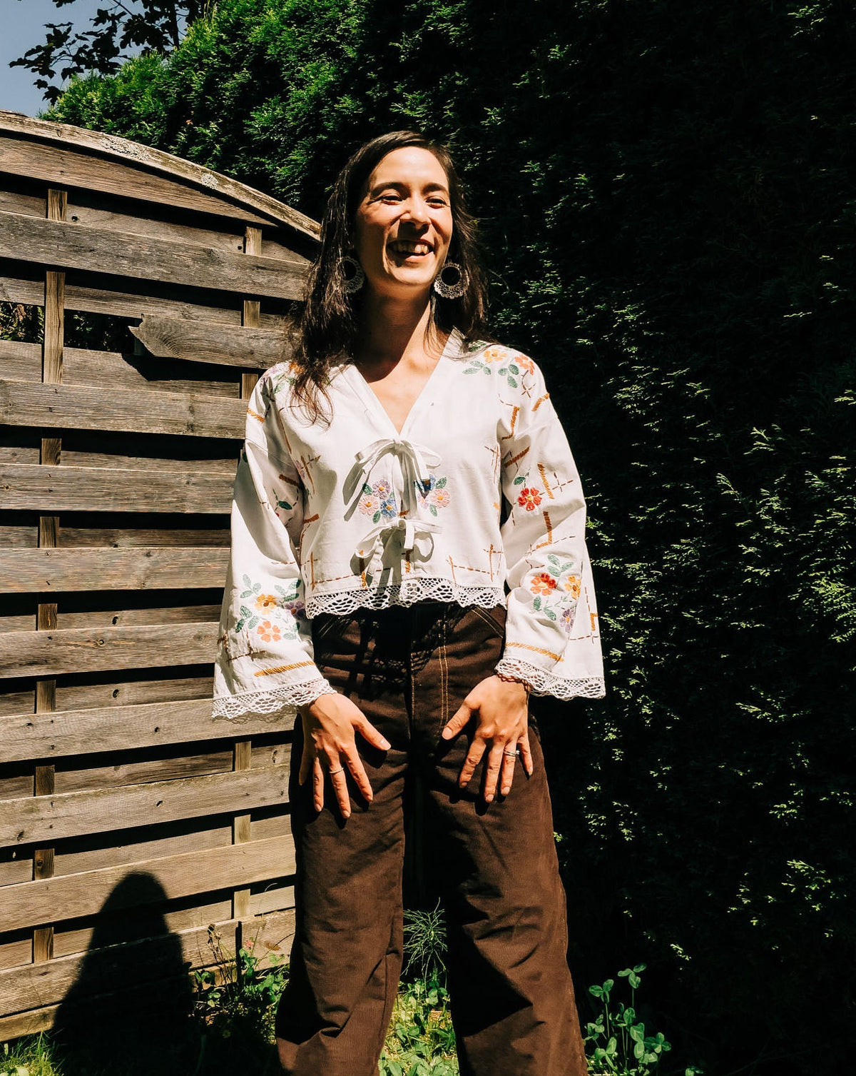Person wearing a white embroidered blouse and brown pants standing outdoors with greenery in the background