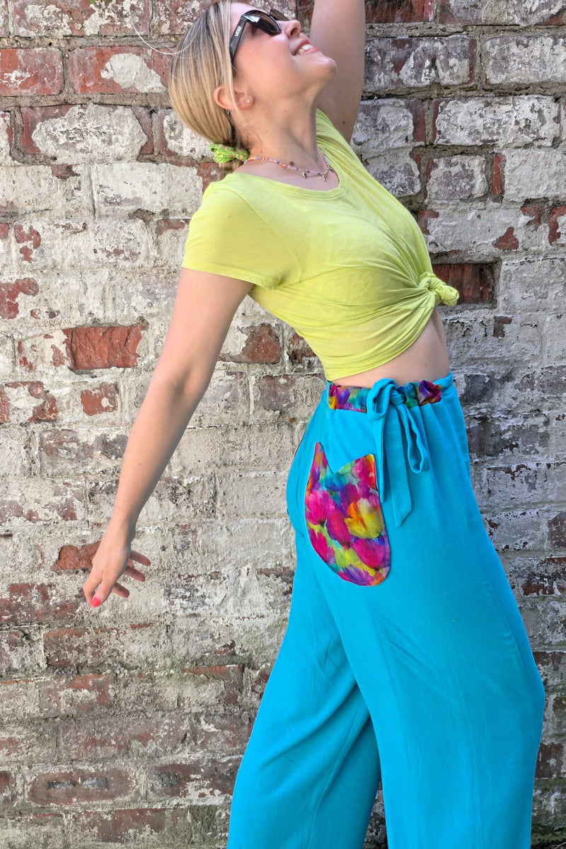A woman wearing bright turquoise wide-leg pants with a colorful tie-dye heart patch on the thigh, a yellow-green sleeveless crop top, and sunglasses, posing against a weathered brick wall.