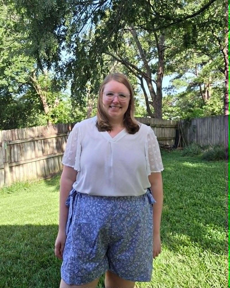 A woman standing outdoors in a grassy backyard wearing a white short-sleeve blouse and blue floral print shorts.