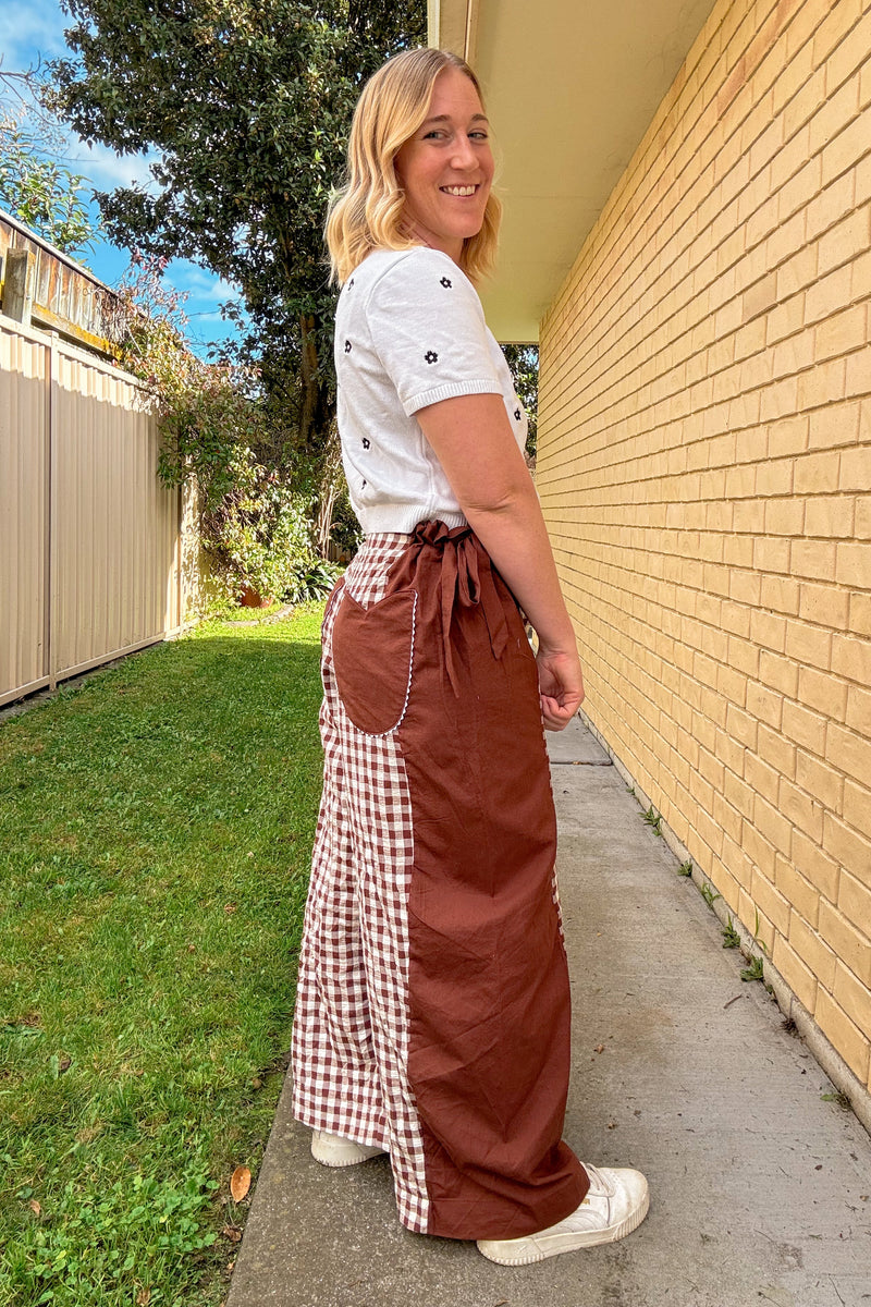 A woman wearing a brown apron-style maxi skirt with a white and red gingham side panel and a white t-shirt, standing outdoors on a concrete path next to a yellow brick wall.