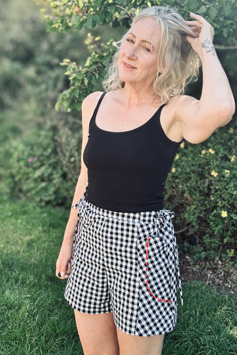 A woman standing outdoors in front of green foliage, wearing a black sleeveless tank top and black-and-white gingham check shorts. The photo appears to be embedded within a mobile app or website interface with status bar and editing tools visible around it.