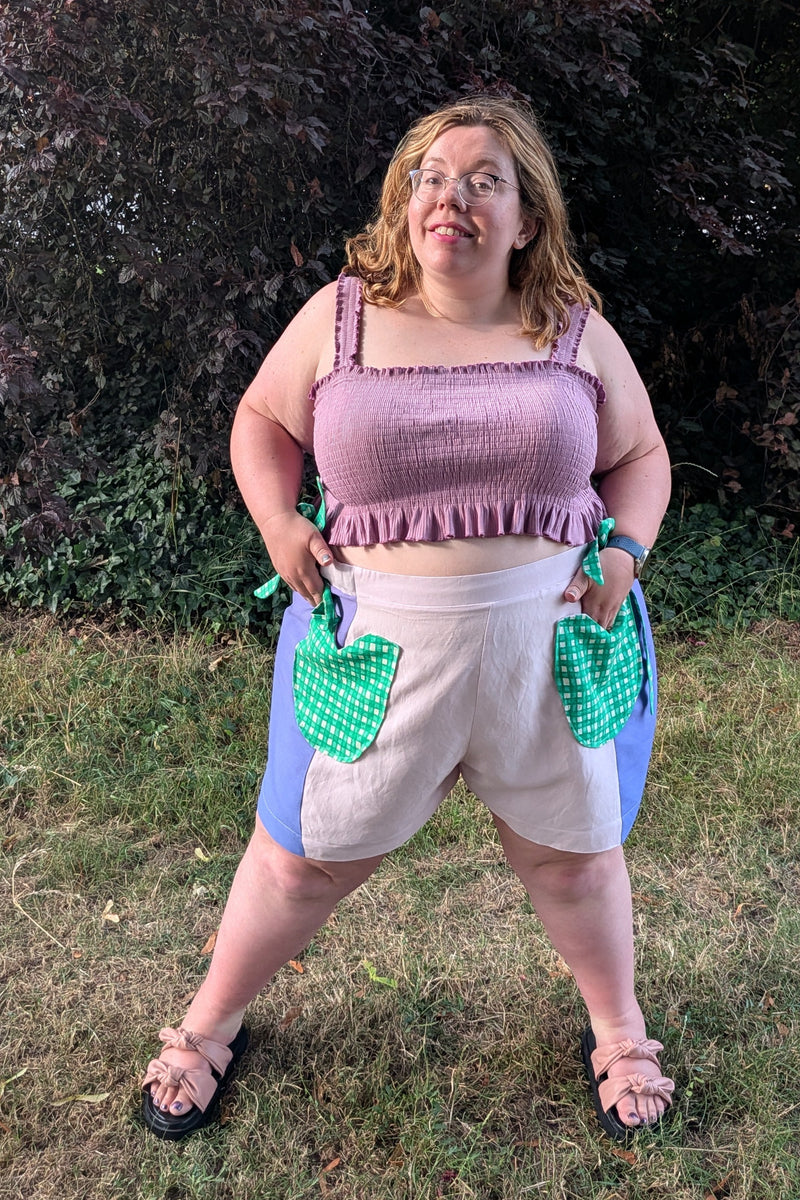 A woman modeling a handmade crochet apron-style top and shorts set outdoors on a grassy lawn with a dark leafy hedge in the background. The set features a lavender-pink ruffled tank top, cream shorts, and green and blue crochet pockets.