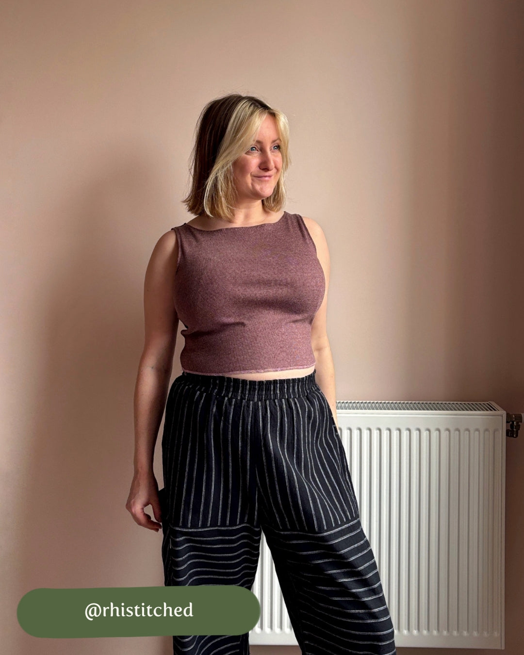 Woman wearing a brown top and black pinstripe pants standing against a beige wall with a radiator.