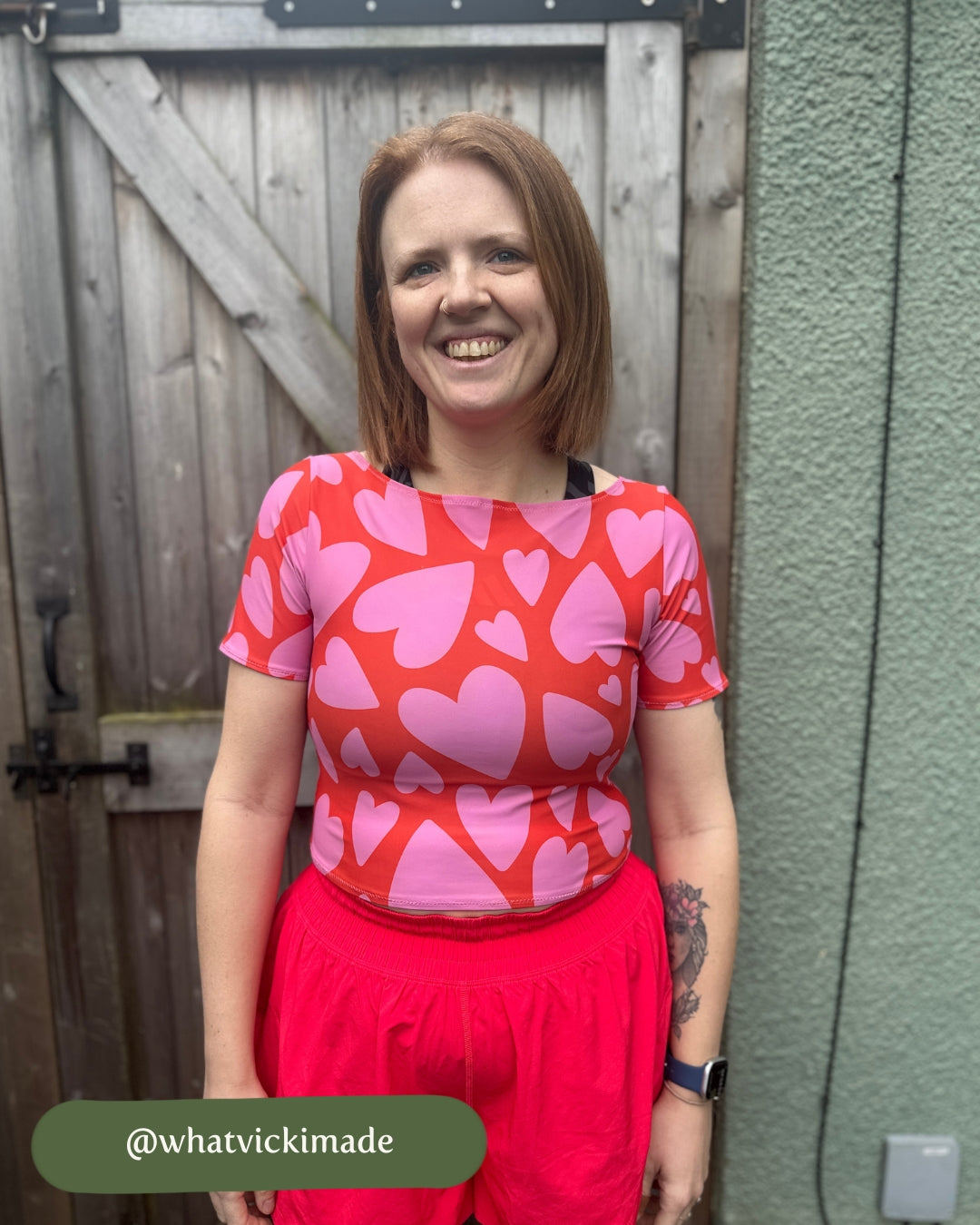 Woman wearing a red and pink heart-patterned top standing against a wooden fence.